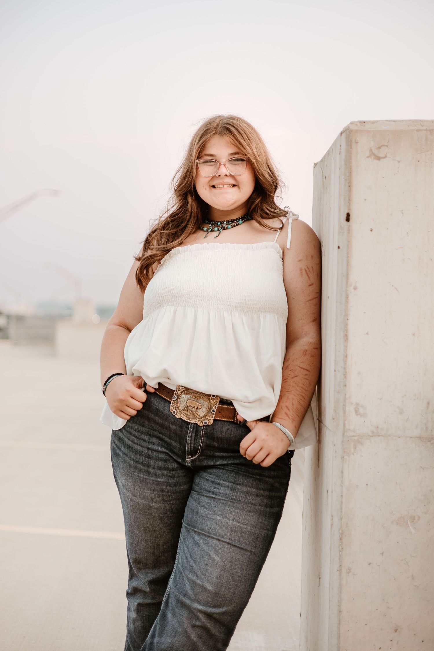 Young woman in white tank top, jeans, and decorative belt buckle smiles while leaning against a concrete pillar outdoors.