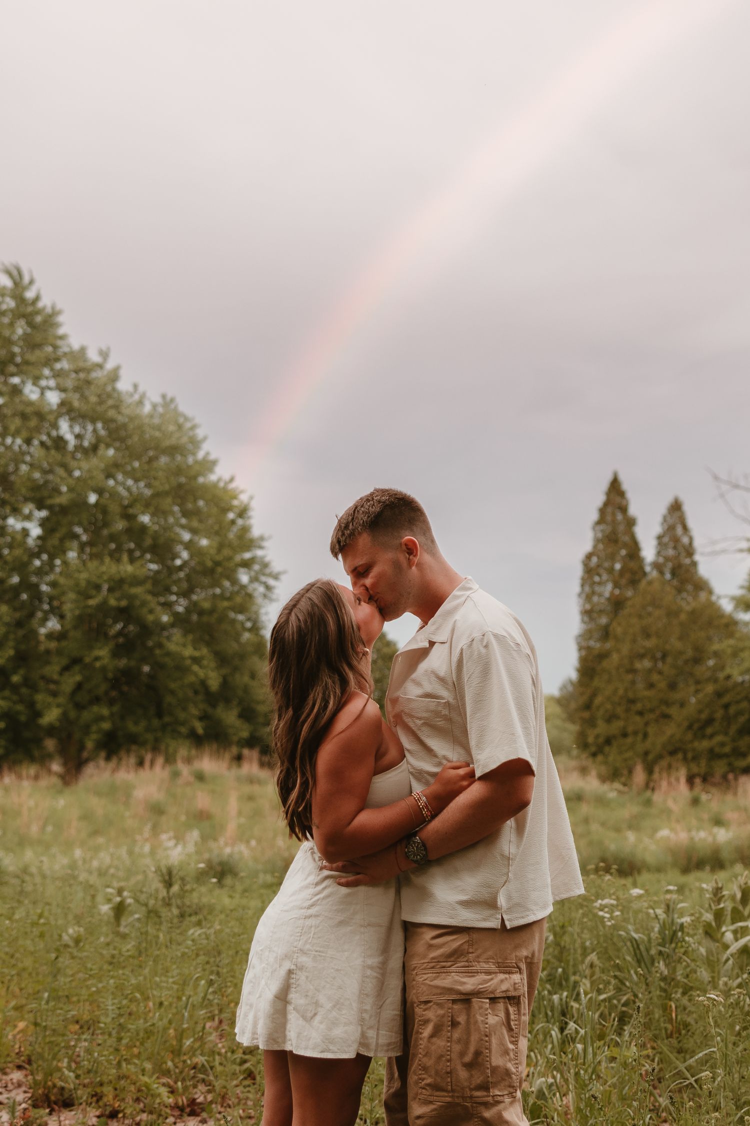 Couple kissing in a lush green field with a rainbow arching across the cloudy sky behind them.