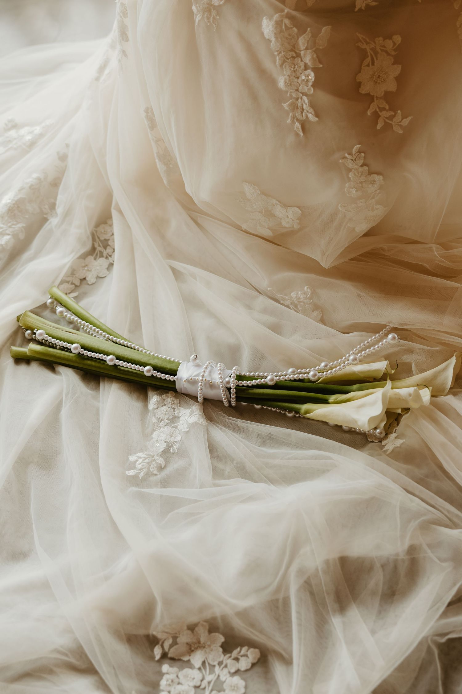 Elegant white calla lily bridal bouquet tied with pearl chain ribbon resting on lace wedding dress fabric.