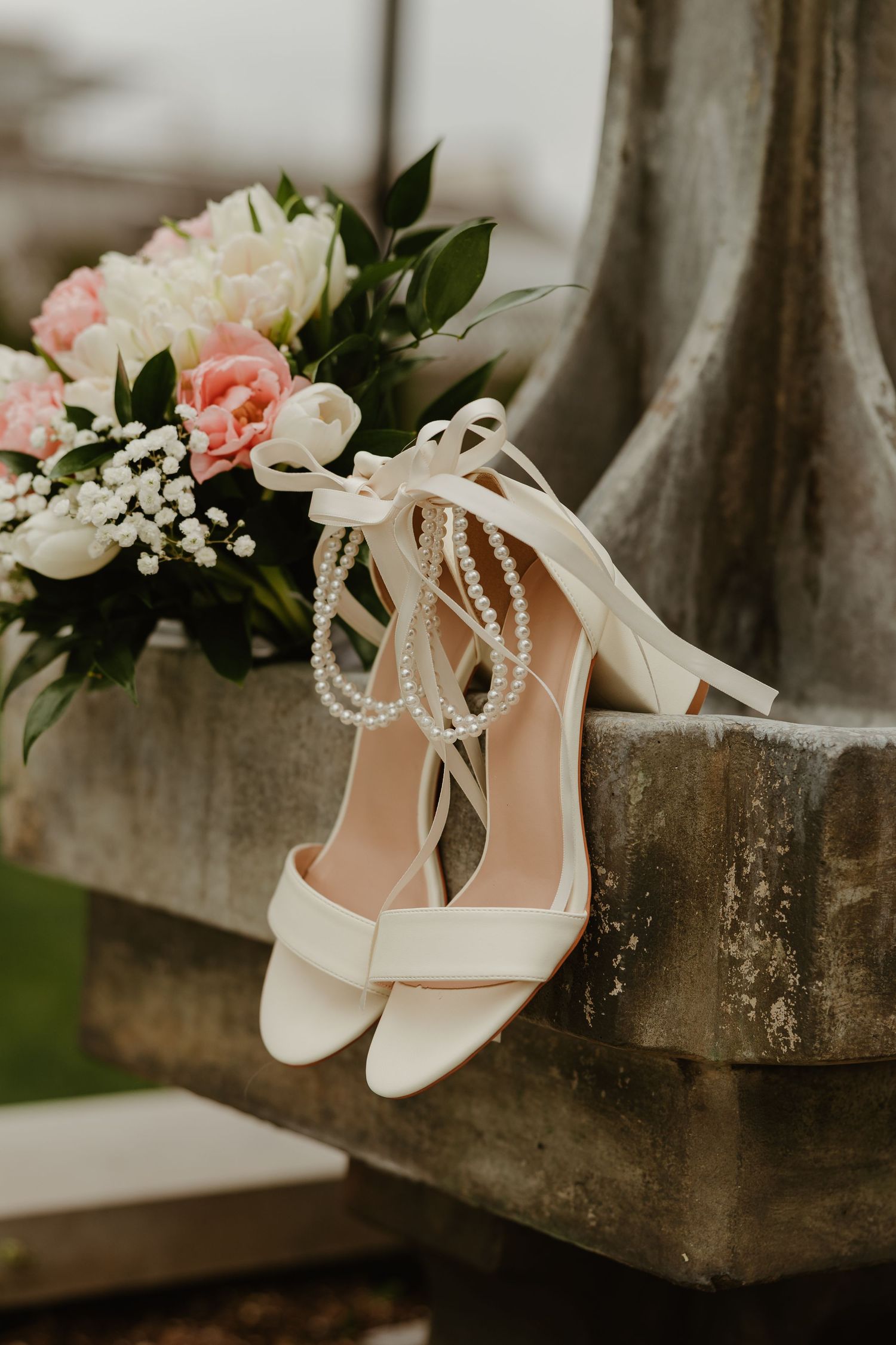 White bridal heels with pearl ankle straps and ribbon bows displayed on stone ledge beside pink and white wedding bouquet.