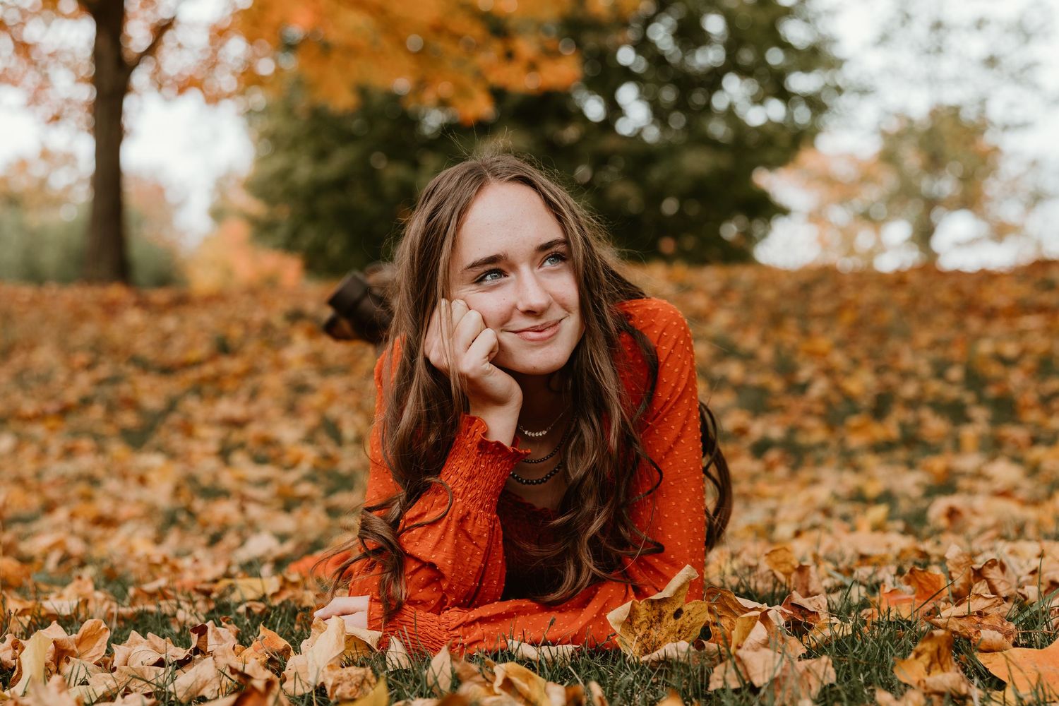 Young woman in orange dress lying on autumn leaves in a park, smiling warmly at camera on a fall day.