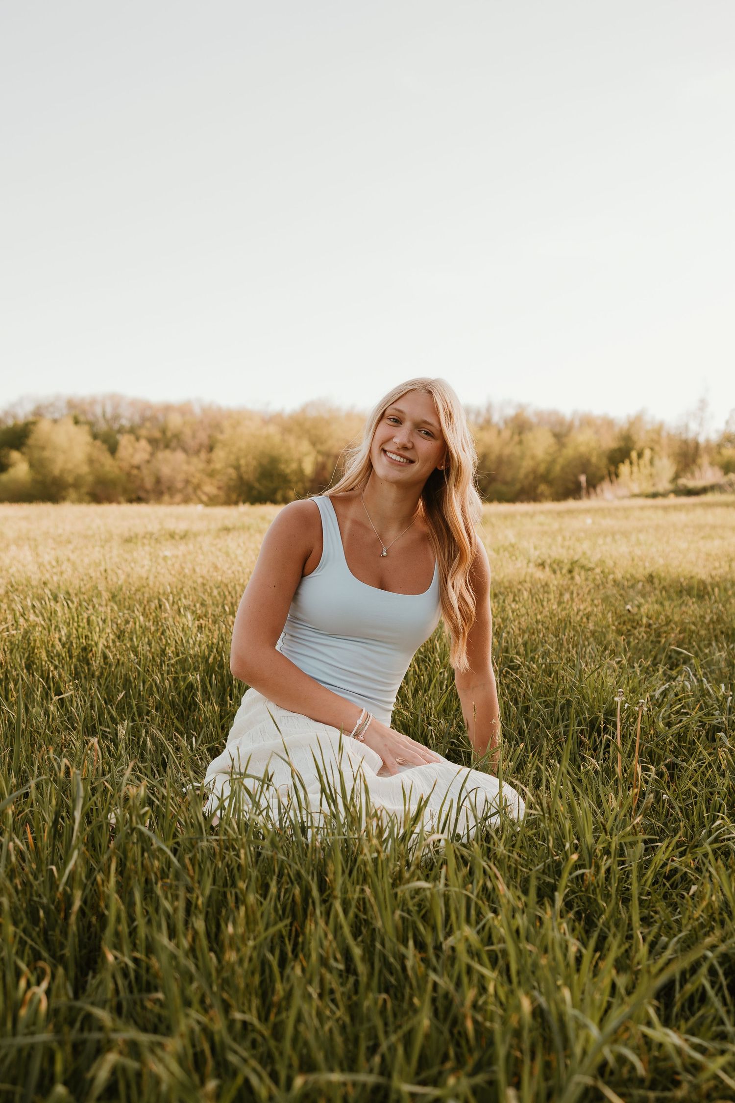 Smiling blonde woman in white skirt and light blue tank top sitting in green grass field with trees in background.