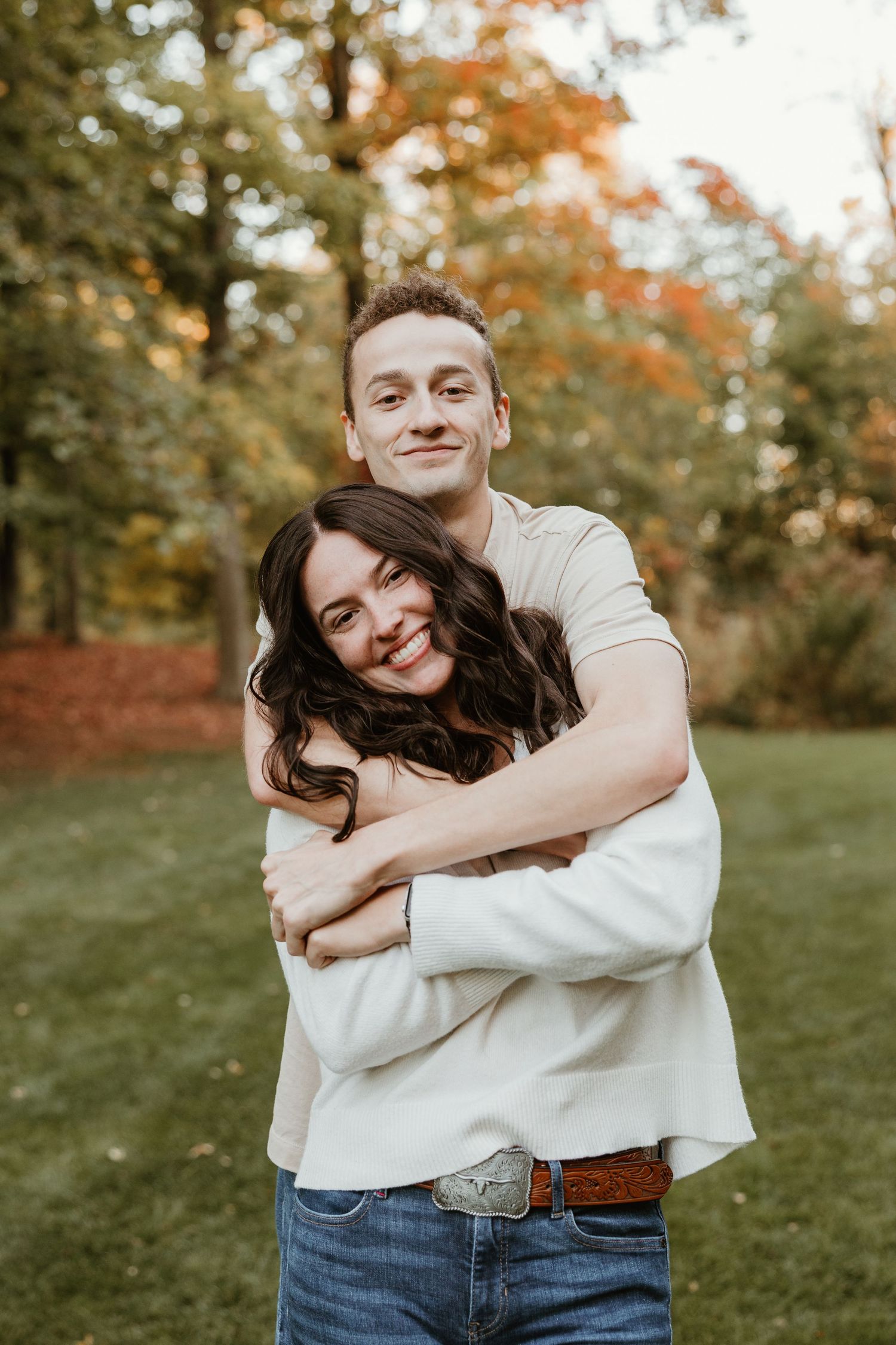 A couple embraces warmly outdoors in autumn, smiling joyfully with colorful fall foliage in the background.