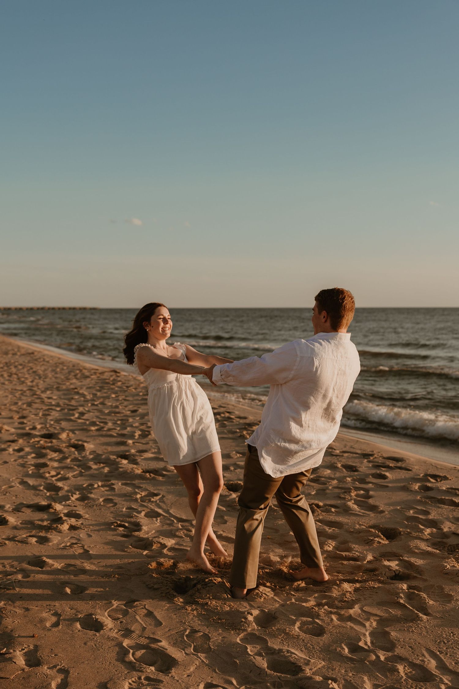 A couple dances barefoot on a sandy beach at sunset, holding hands in white outfits with ocean waves behind them.