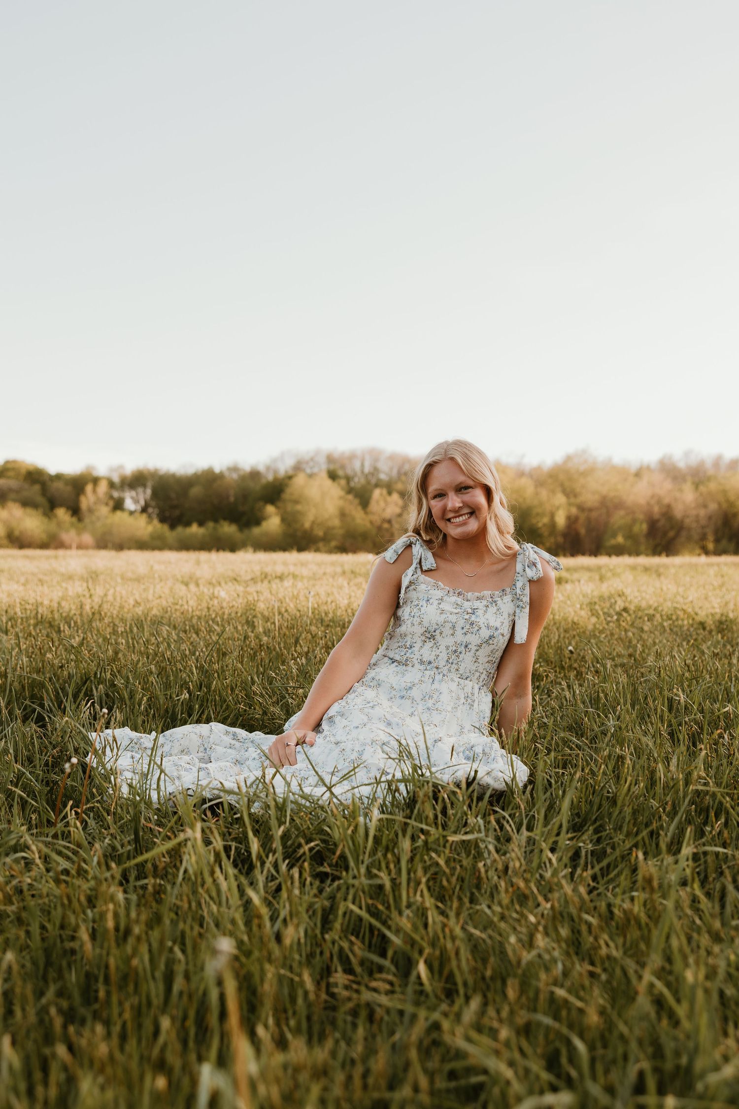 Smiling blonde woman in a floral dress sitting in a lush green field at golden hour.