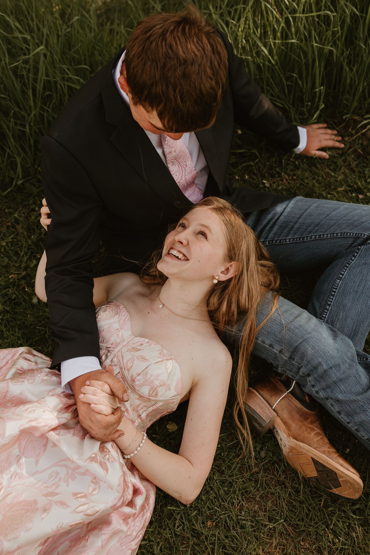 A young couple in formal wear laughing together on green grass, she in a pink gown and cowboy boots.
