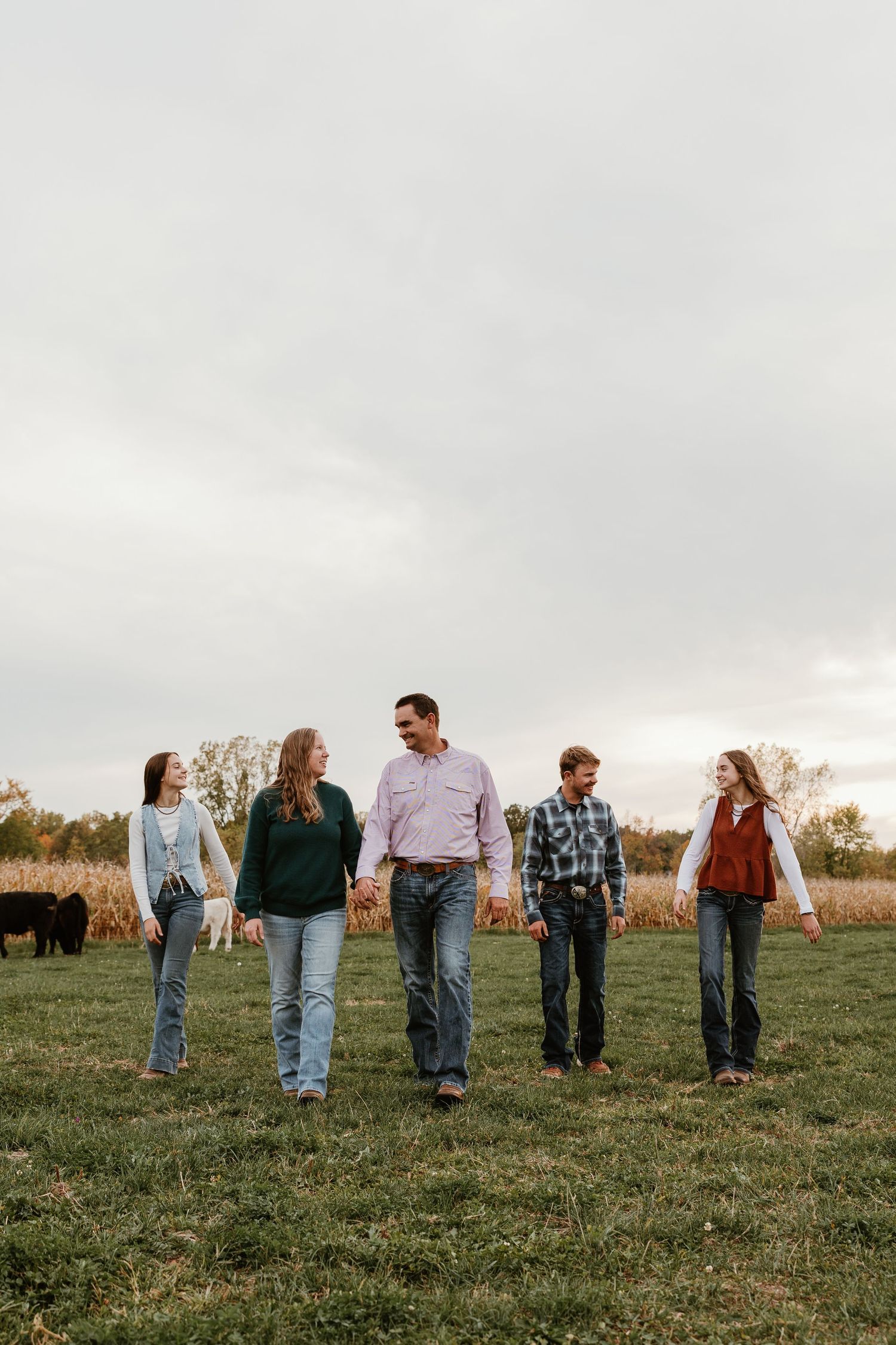 A family of five walks hand-in-hand through a green pasture with cattle and cornfields in the background.