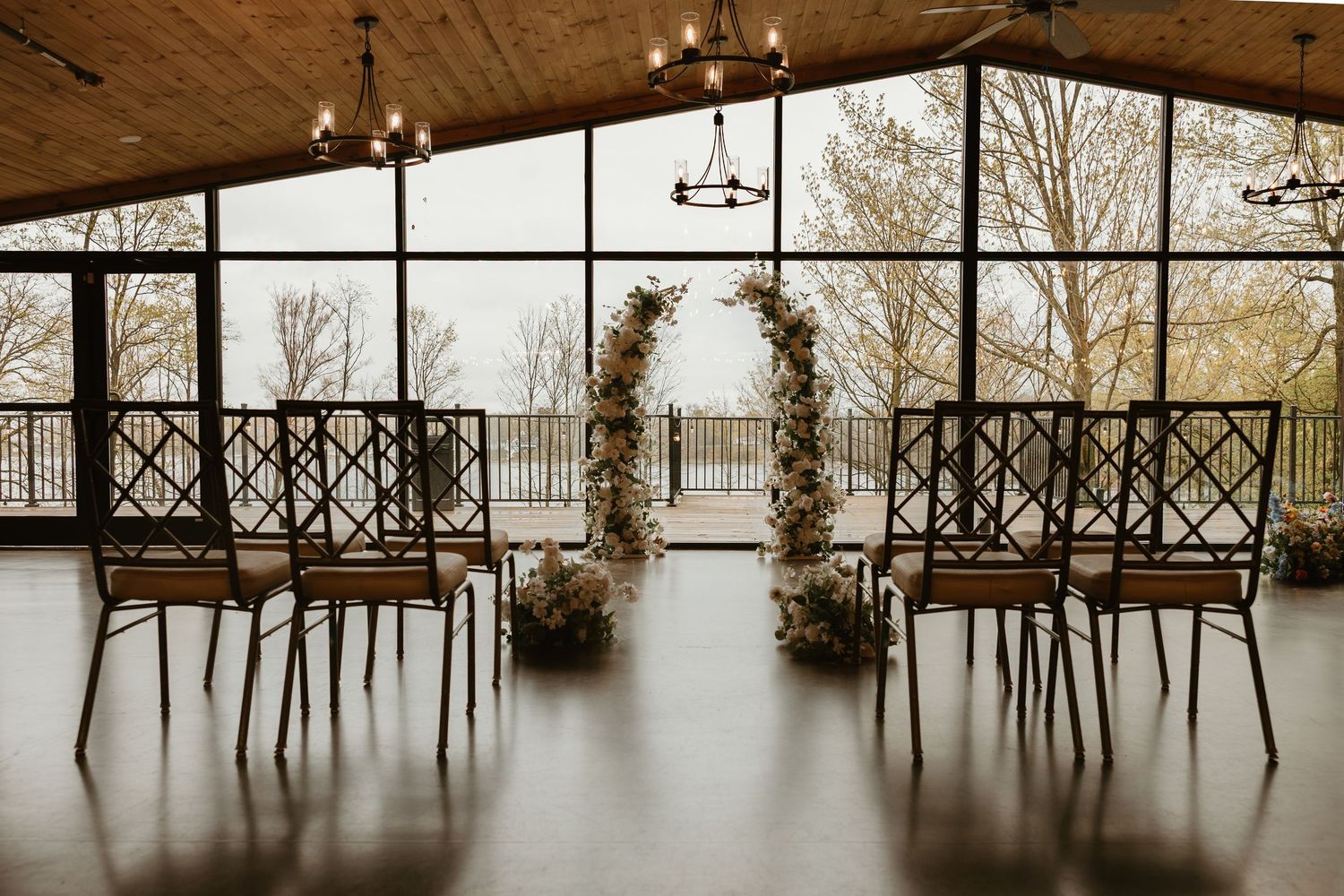 Elegant indoor wedding ceremony setup with floral arch, dark chairs, string lights, and large windows overlooking trees.