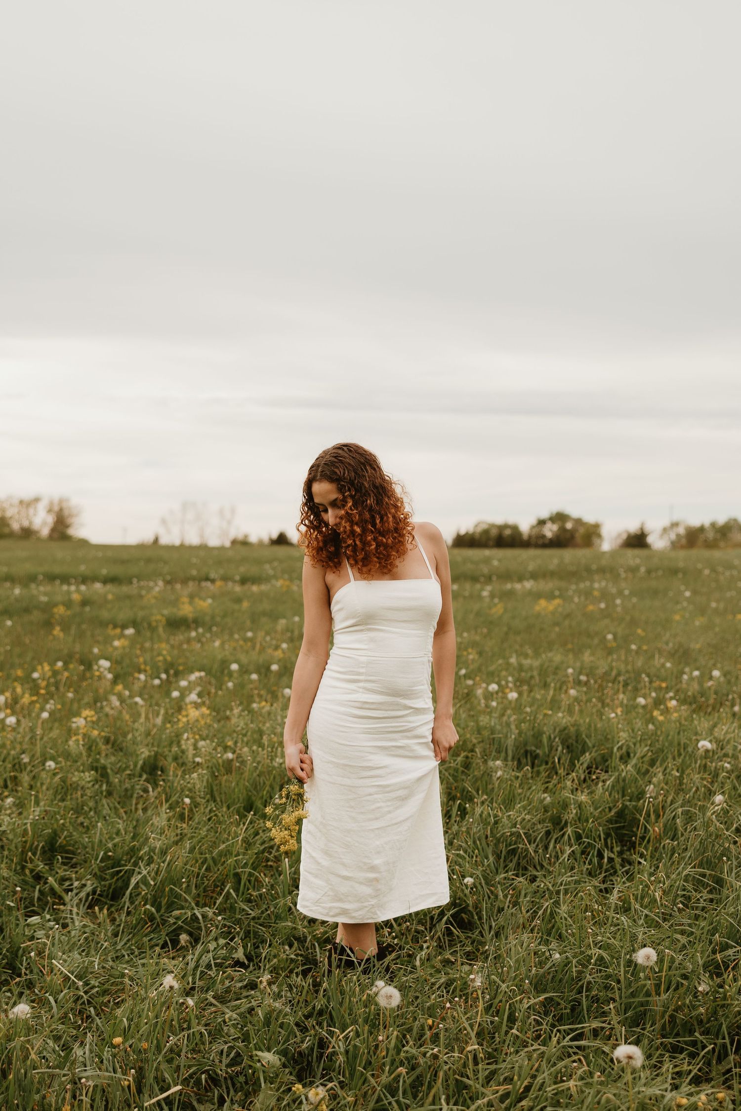 Woman in white midi dress standing in green field with wildflowers under overcast sky.