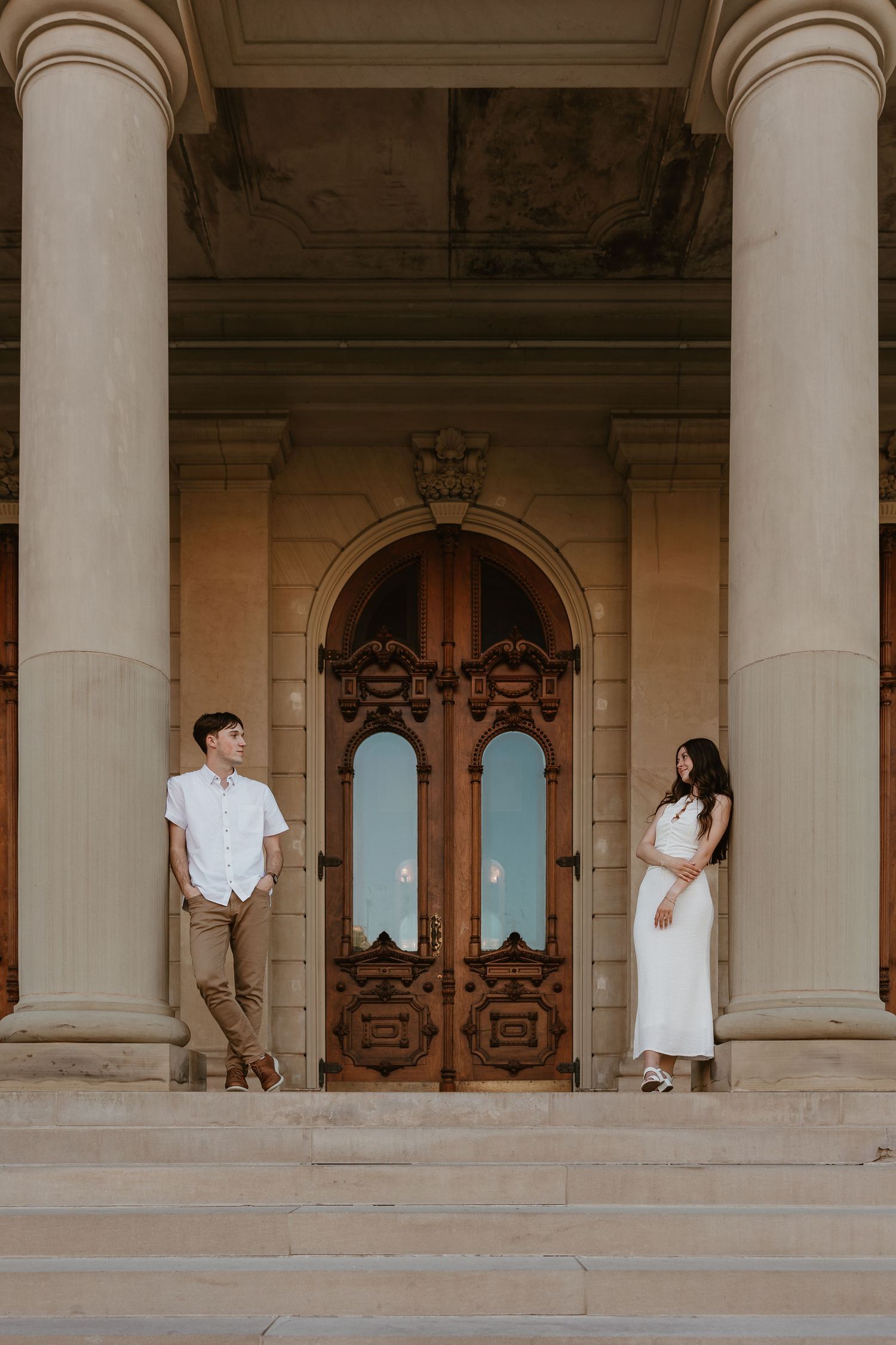 Couple standing apart on steps of elegant building with ornate wooden doors and tall columns.