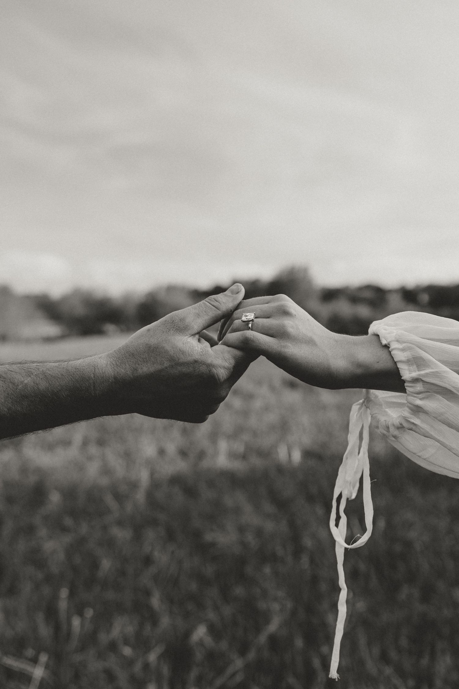 Black and white photo of two hands gently touching outdoors, one wearing a ring, with a blurred field background.