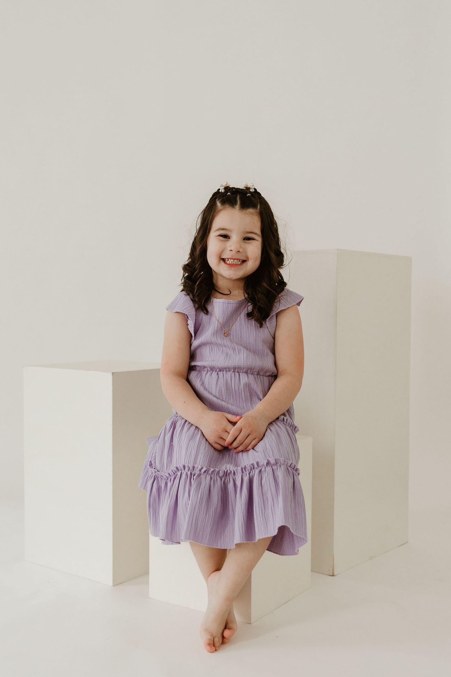 Young girl in lavender ruffle dress smiling, seated on white cube in bright studio setting.