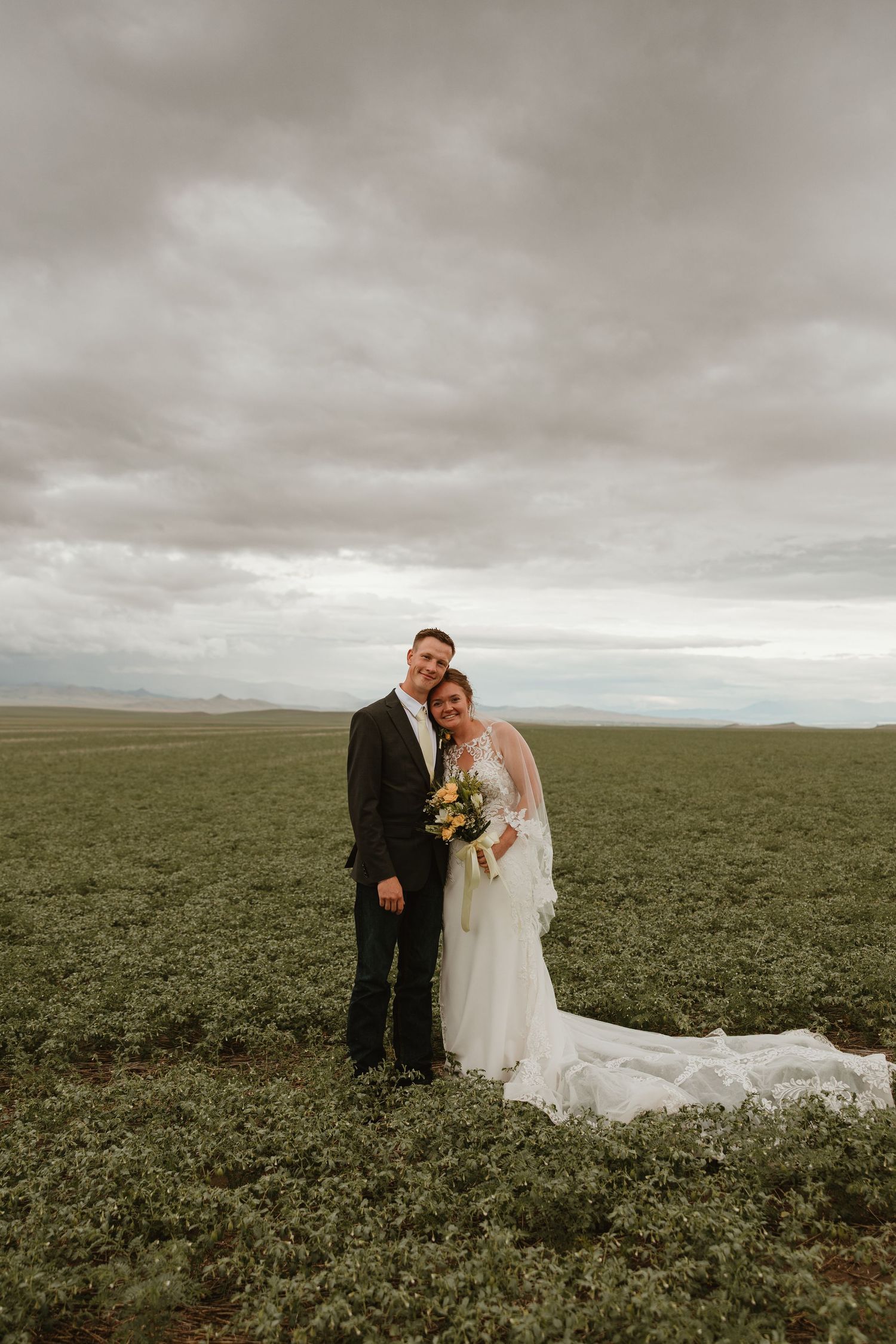 Bride and groom pose in a vast green field under cloudy skies, bride holding a bouquet in a flowing white gown.