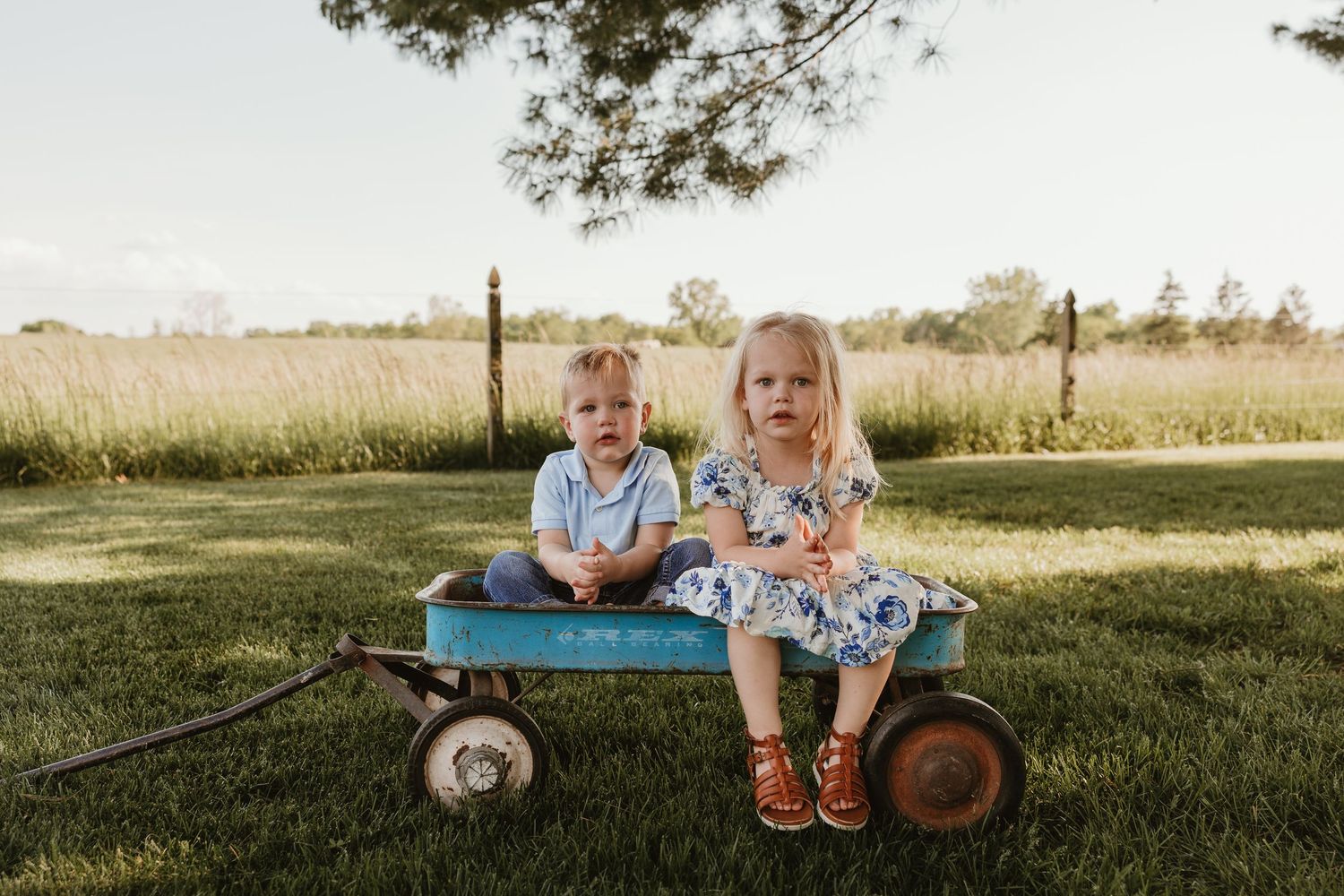 Two young children sitting on a vintage teal wagon on green grass in a sunny outdoor setting.