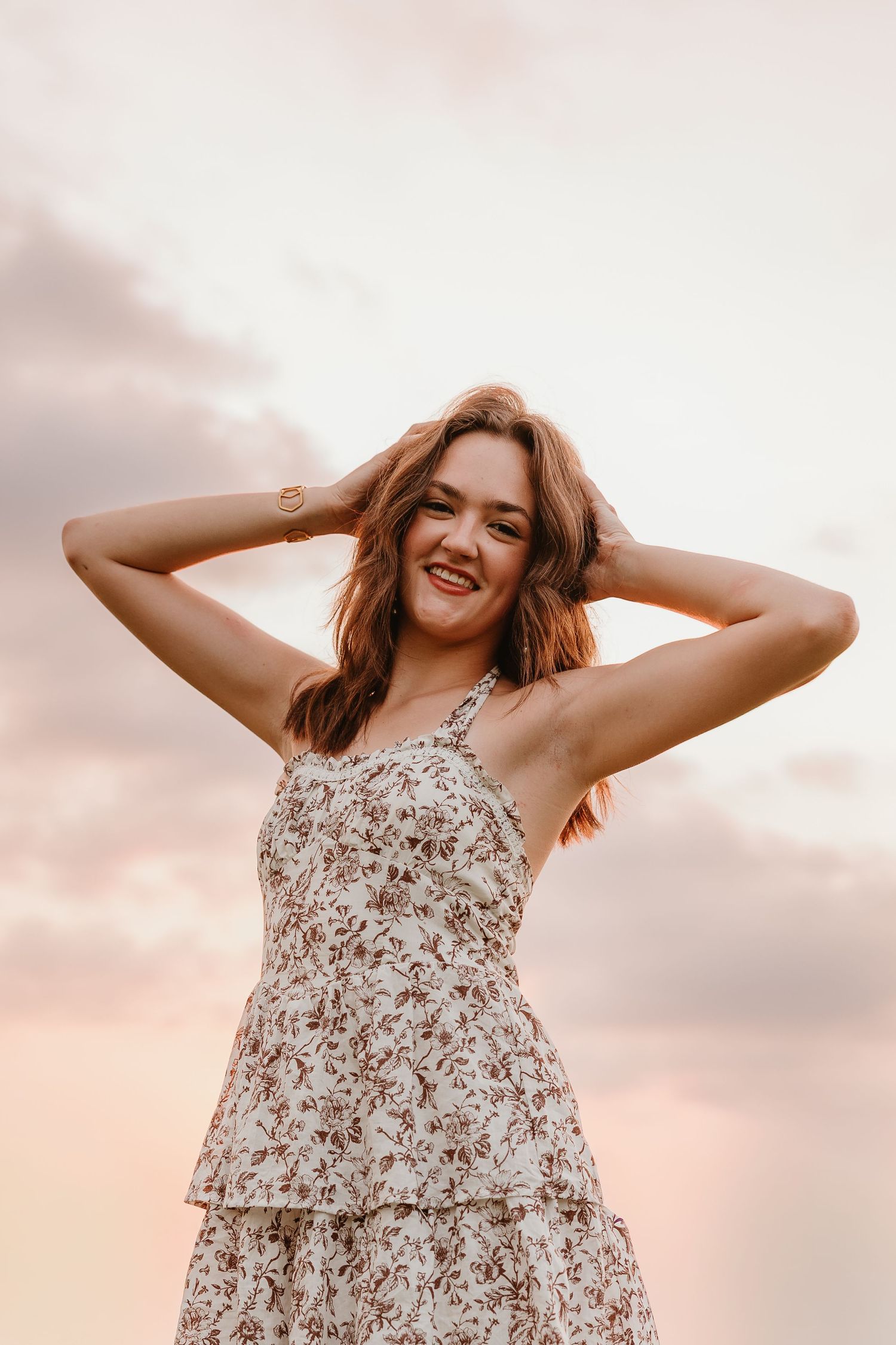 Smiling young woman in floral sundress poses outdoors at sunset with hands in her hair.