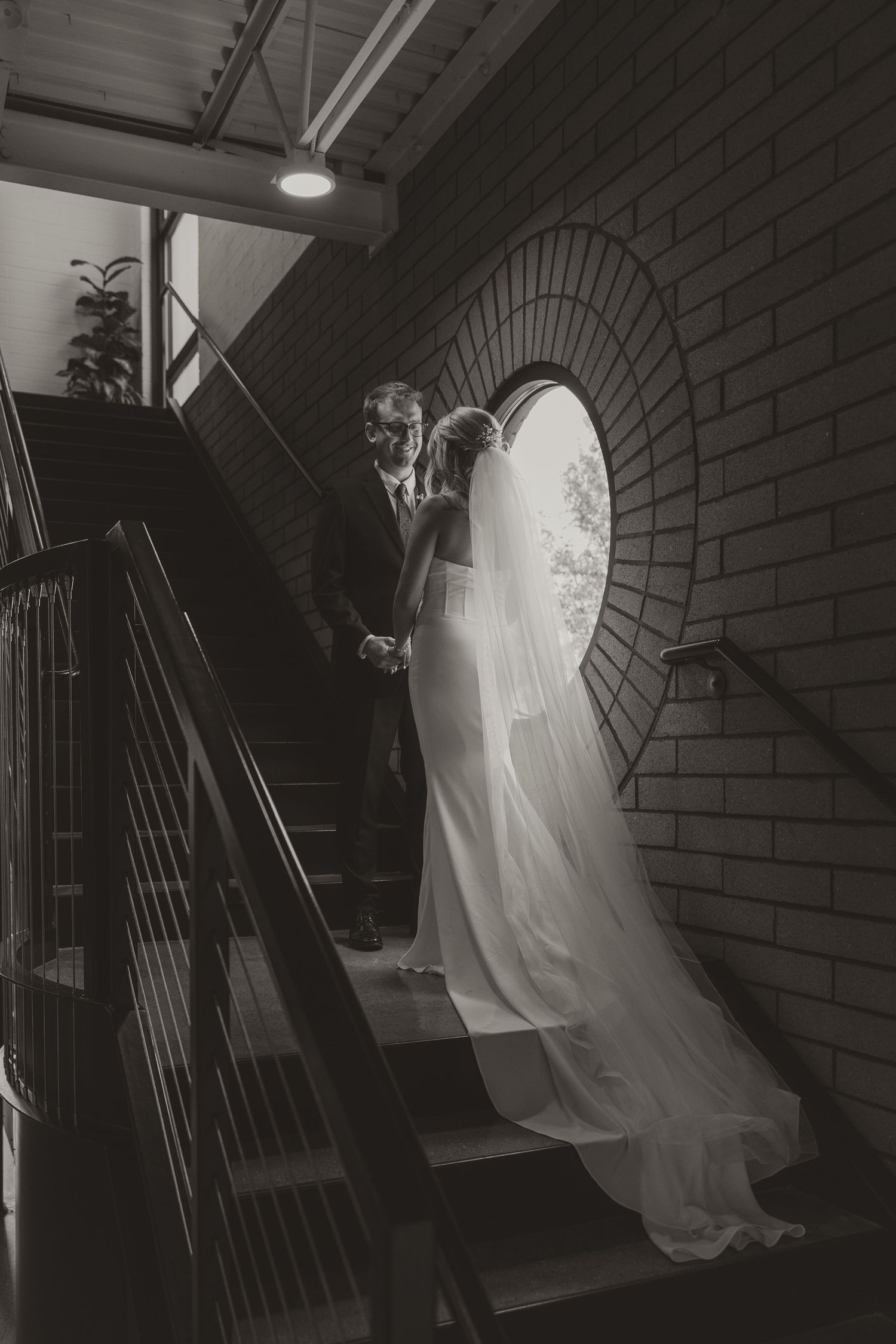 Black and white photo of bride and groom holding hands on staircase beside circular brick window.