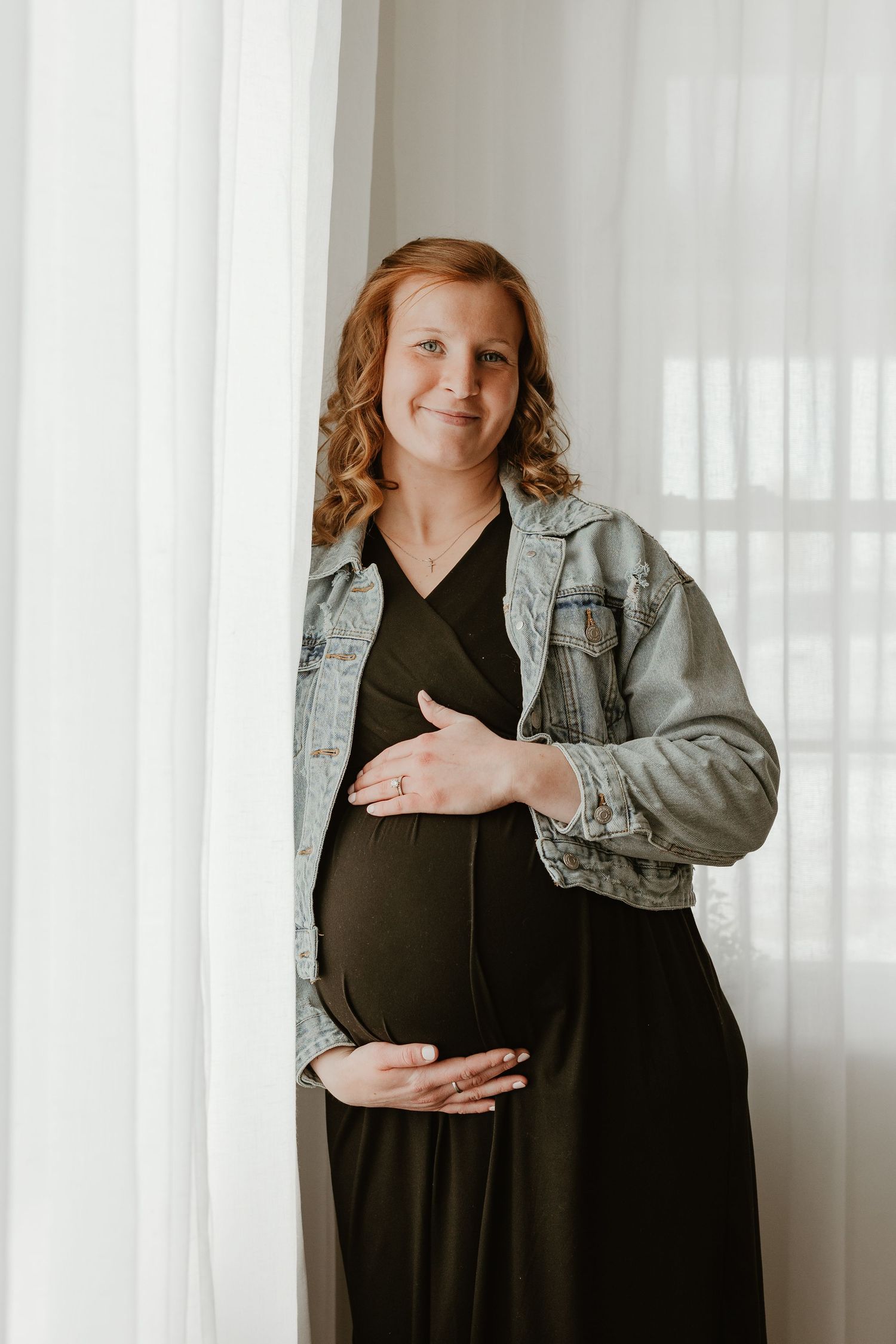 Pregnant woman in black dress and denim jacket cradling baby bump by white curtains.