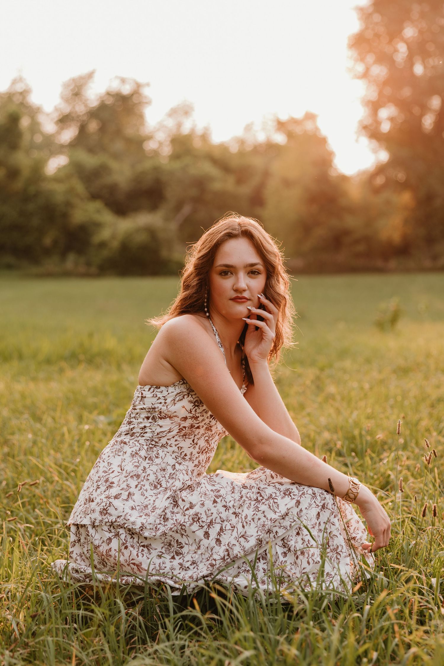 Young woman in floral dress sitting in golden hour sunlit meadow, posing gracefully with trees in background.