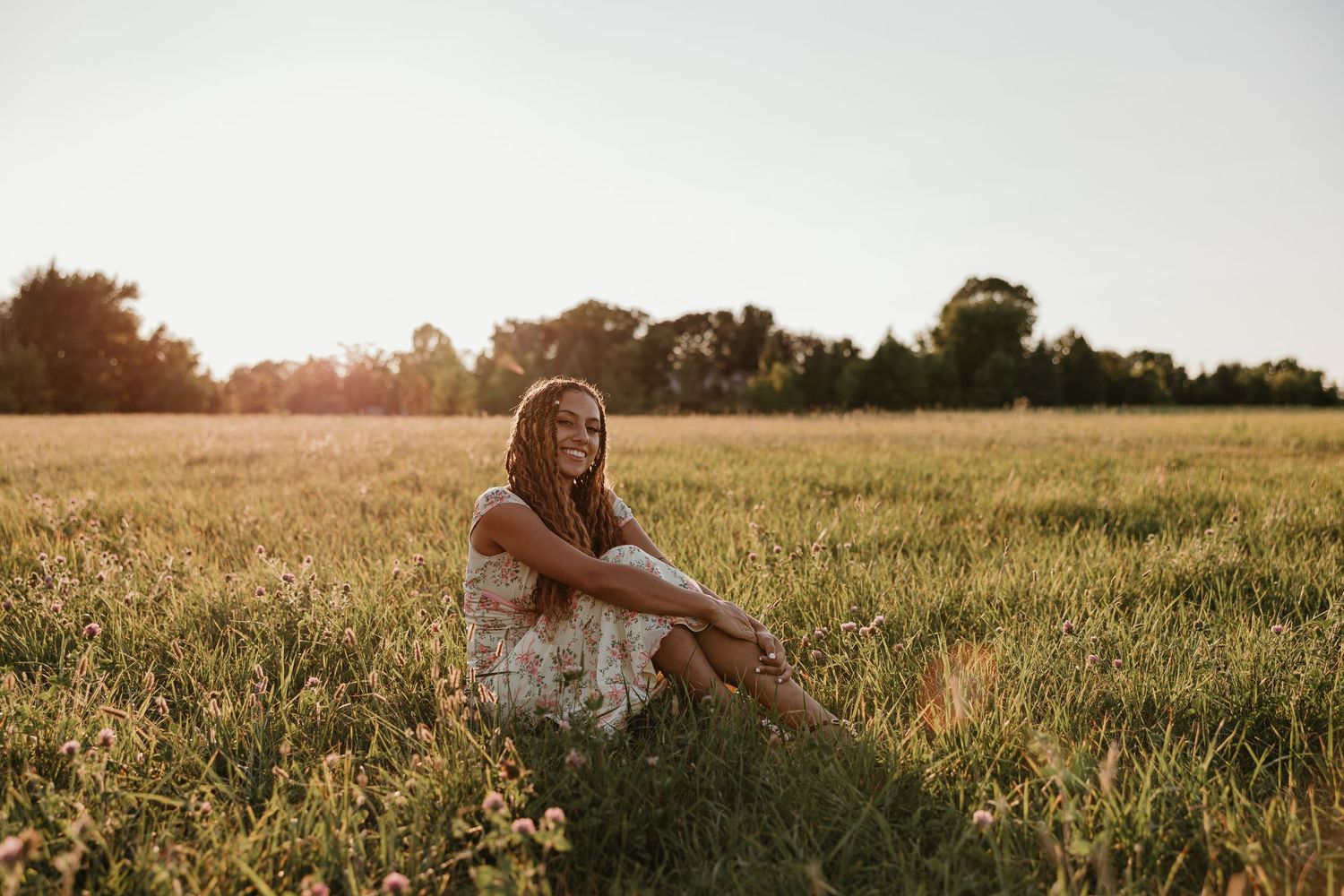 Young woman in floral dress sitting in sunlit meadow at golden hour with trees in background.