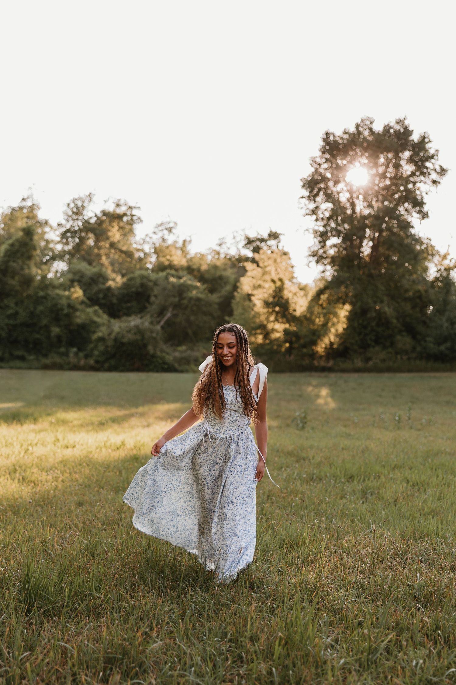 Young woman in white floral dress twirling in sunlit green meadow at golden hour with trees in background.