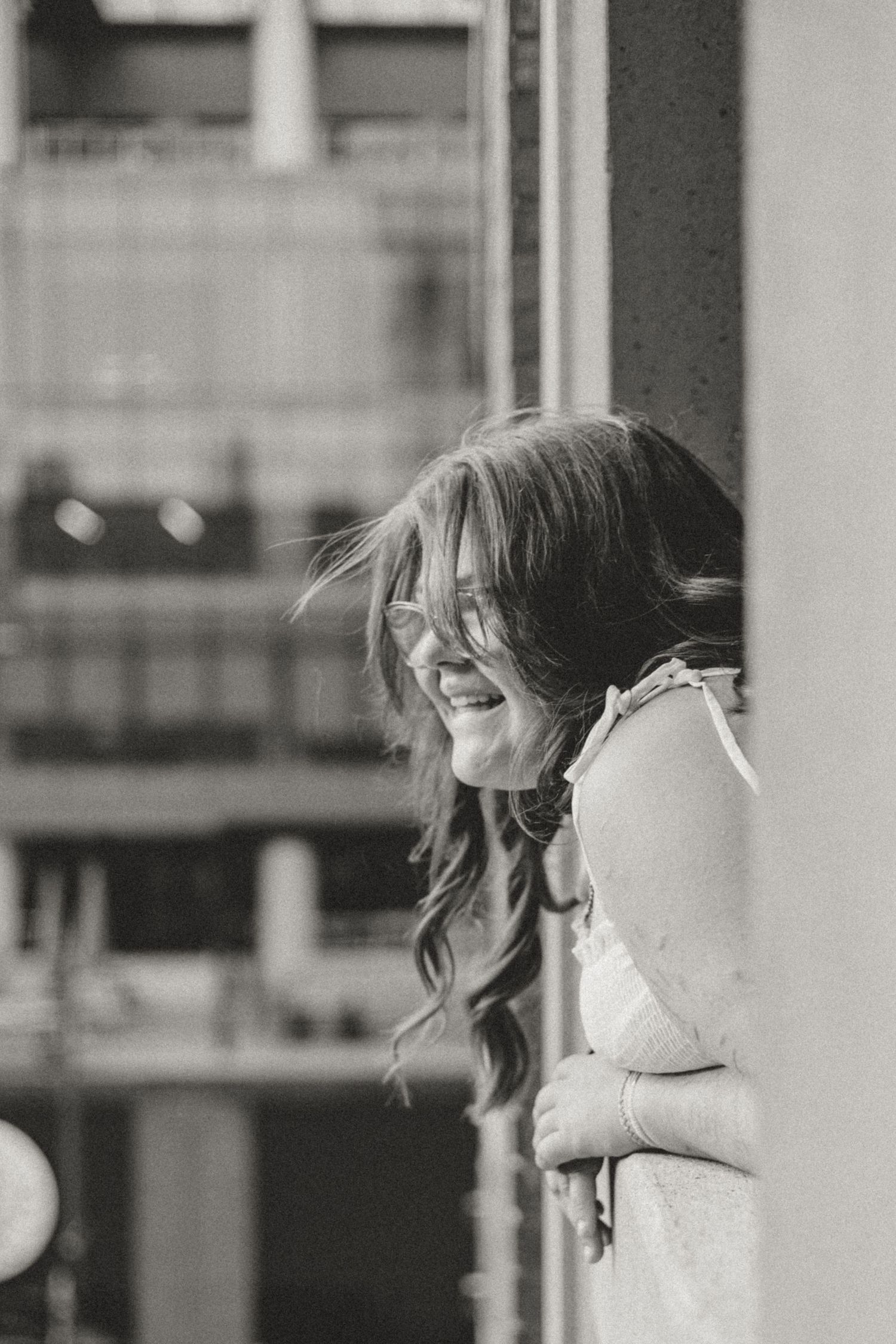 Black and white photo of a young woman laughing outdoors, hair blowing in the wind, leaning against a wall.