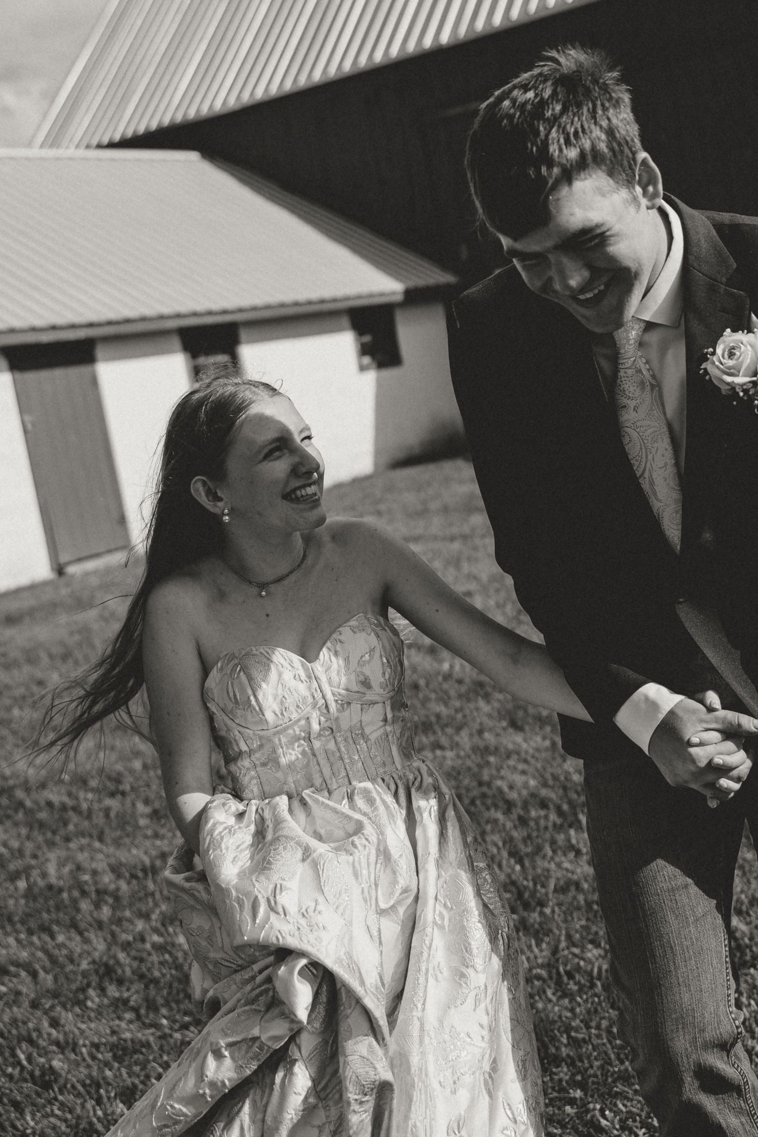 Black and white photo of a joyful bride and groom holding hands and laughing outside a rustic barn on their wedding day.