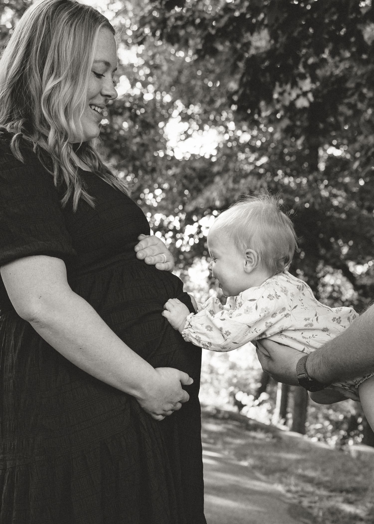 Black and white photo of a pregnant woman smiling as a baby leans in to kiss her baby bump outdoors.