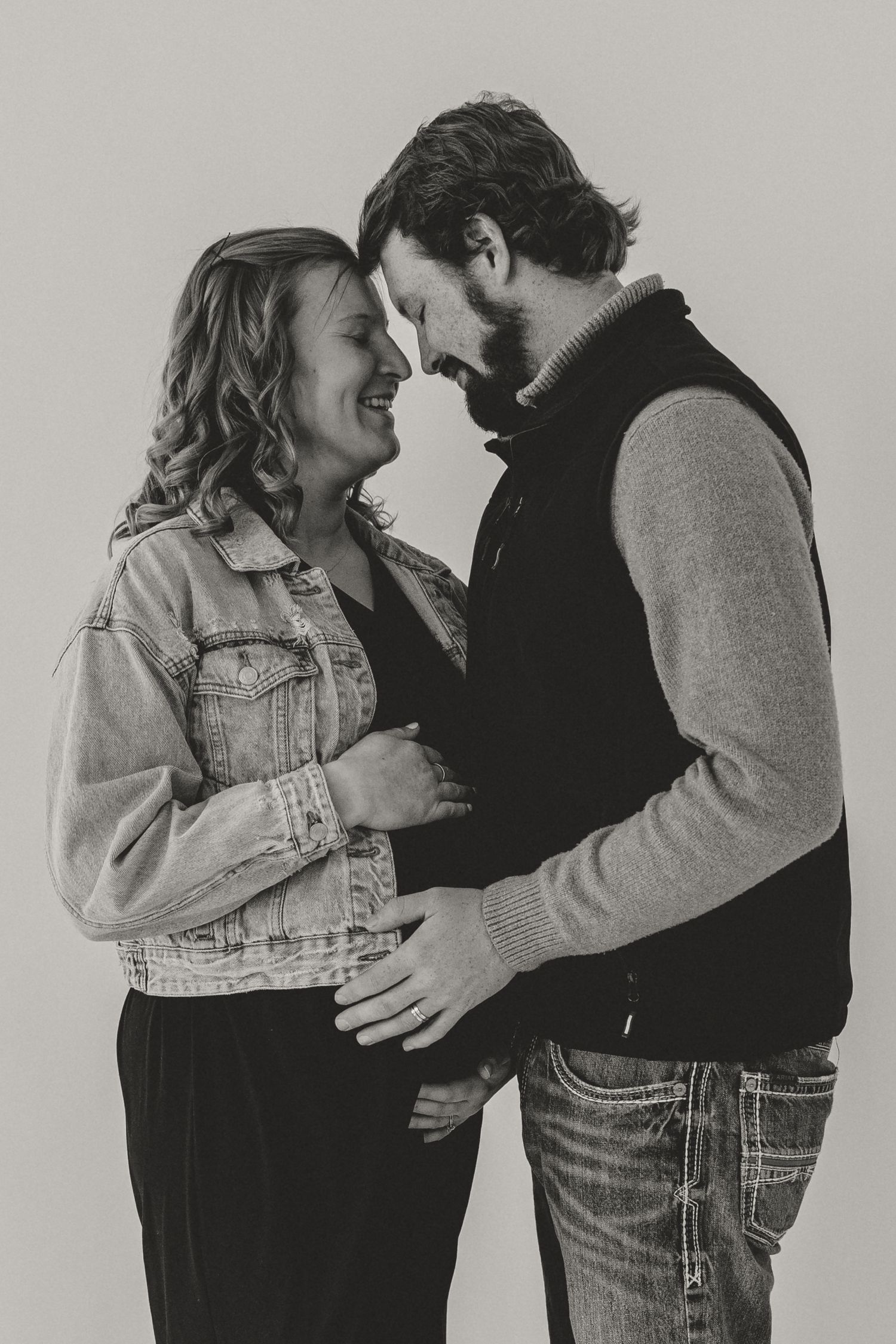 Black and white photo of a smiling couple sharing a tender moment, with hands gently placed on the woman's baby bump.