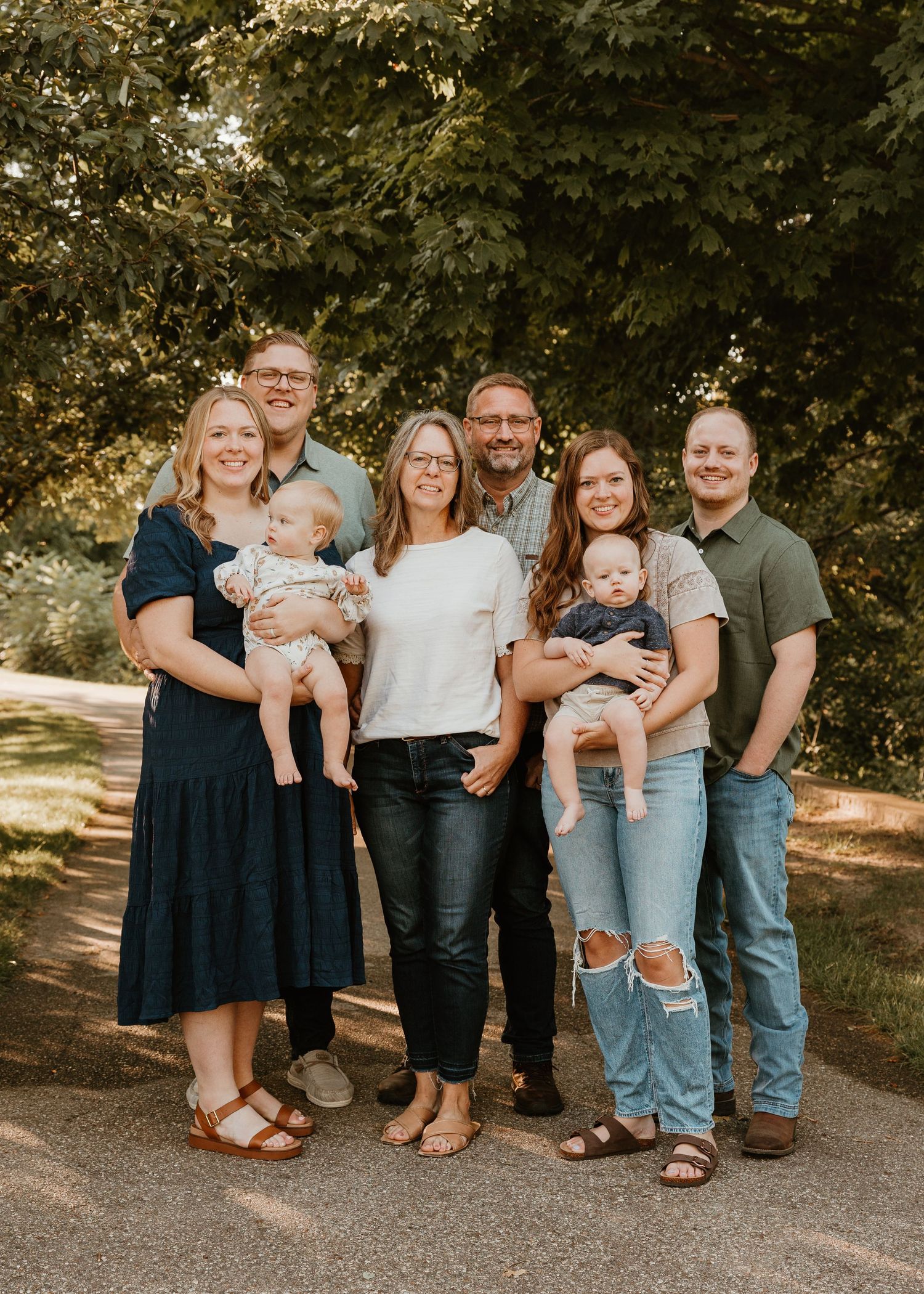Multi-generational family portrait outdoors on a tree-lined path, featuring adults and two babies smiling together.