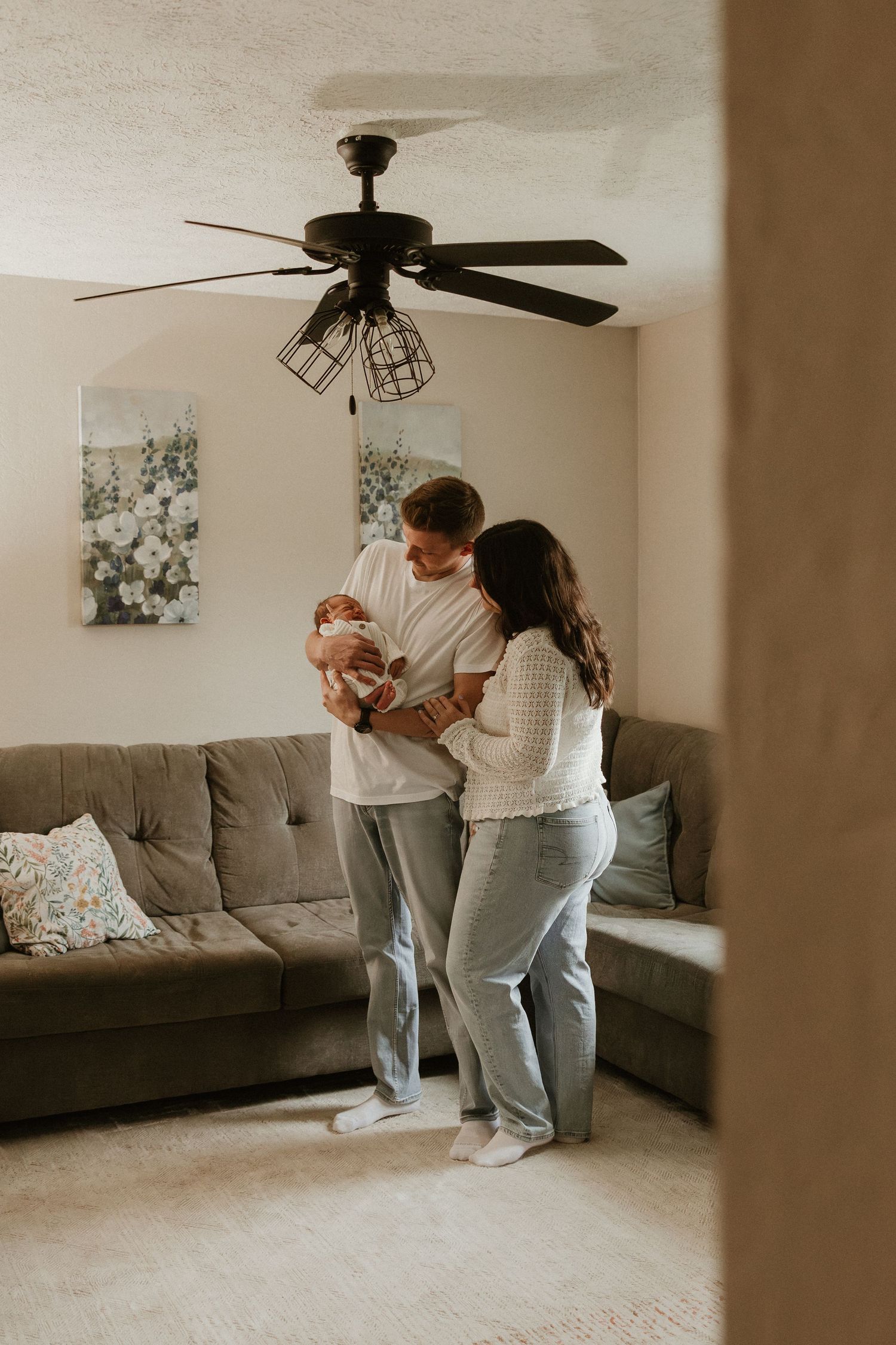 Young couple standing in cozy living room holding their newborn baby under a ceiling fan.