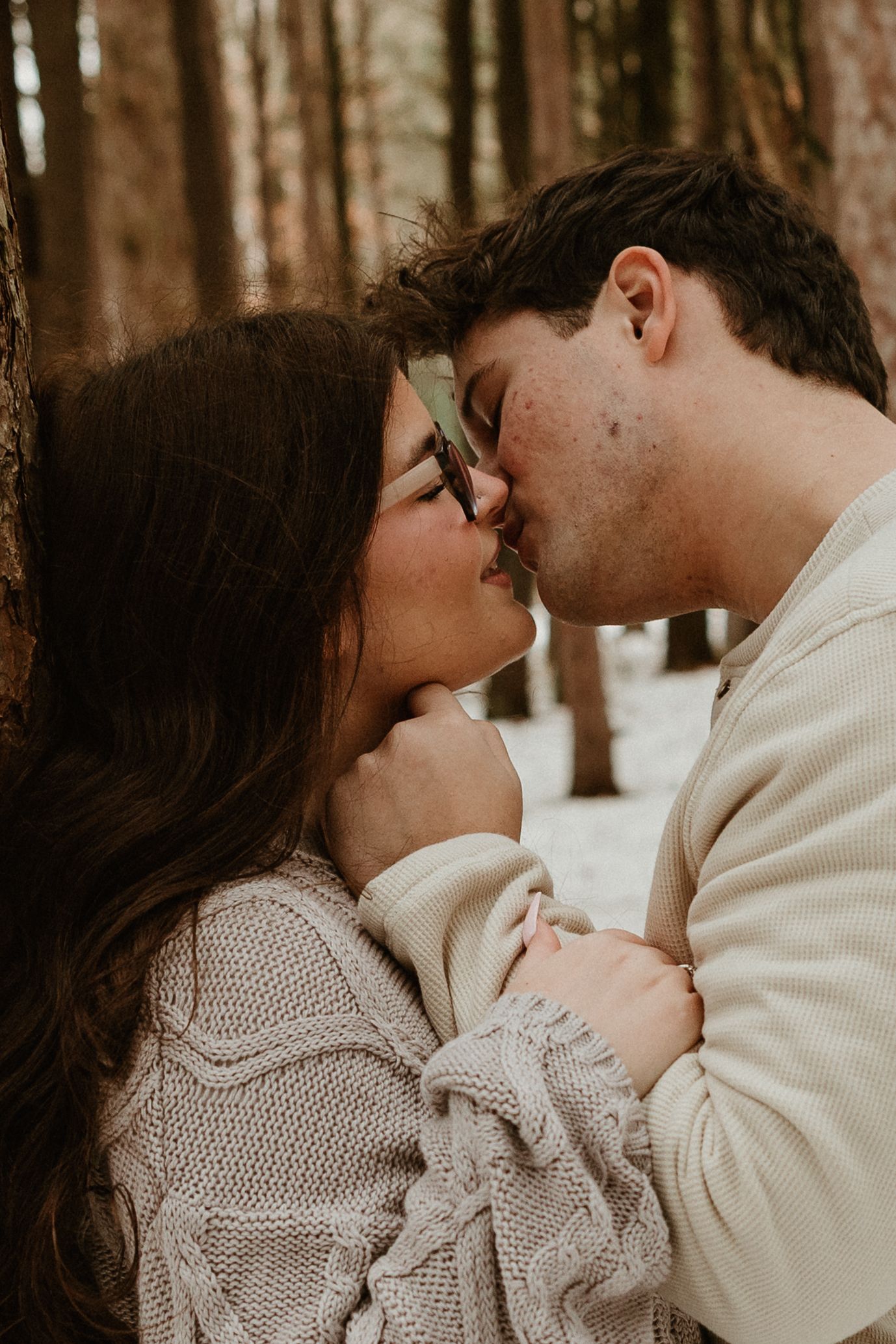 A couple shares a romantic kiss among snow-dusted trees, dressed in cozy cream sweaters during a winter forest photoshoot.