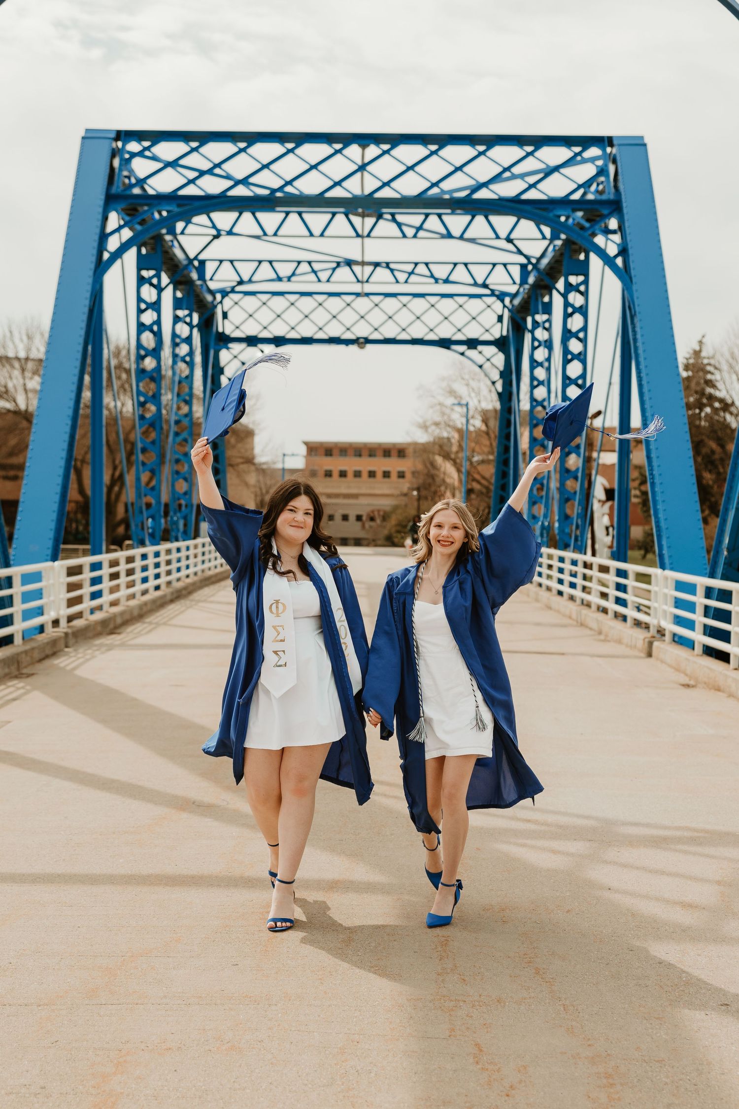 Two women in navy blue graduation gowns and caps celebrate on a blue bridge, wearing white dresses and blue heels.