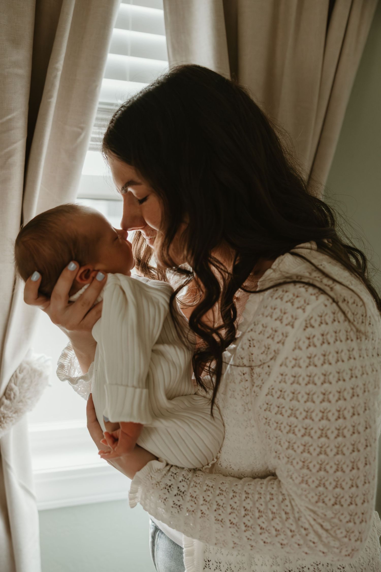 A mother tenderly kisses her newborn baby by a window, both dressed in white lace outfits.