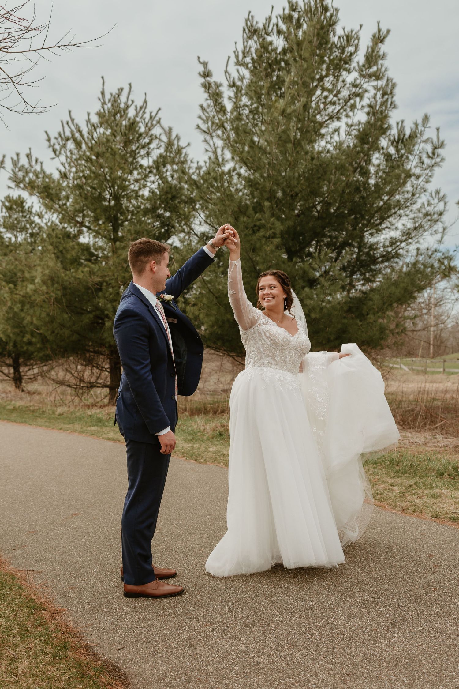 A groom in a navy suit twirls his bride in a white lace gown on a path surrounded by evergreen trees.