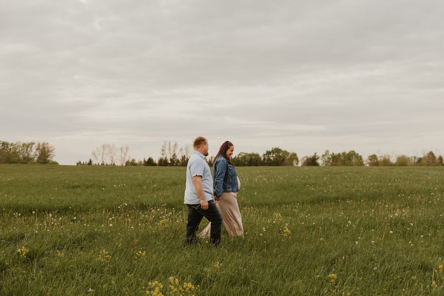 Couple holding hands walking through a lush green field with wildflowers under a cloudy sky.