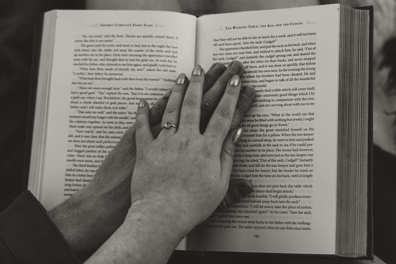 Two hands resting on an open book, one wearing a diamond ring, in a black and white photo.