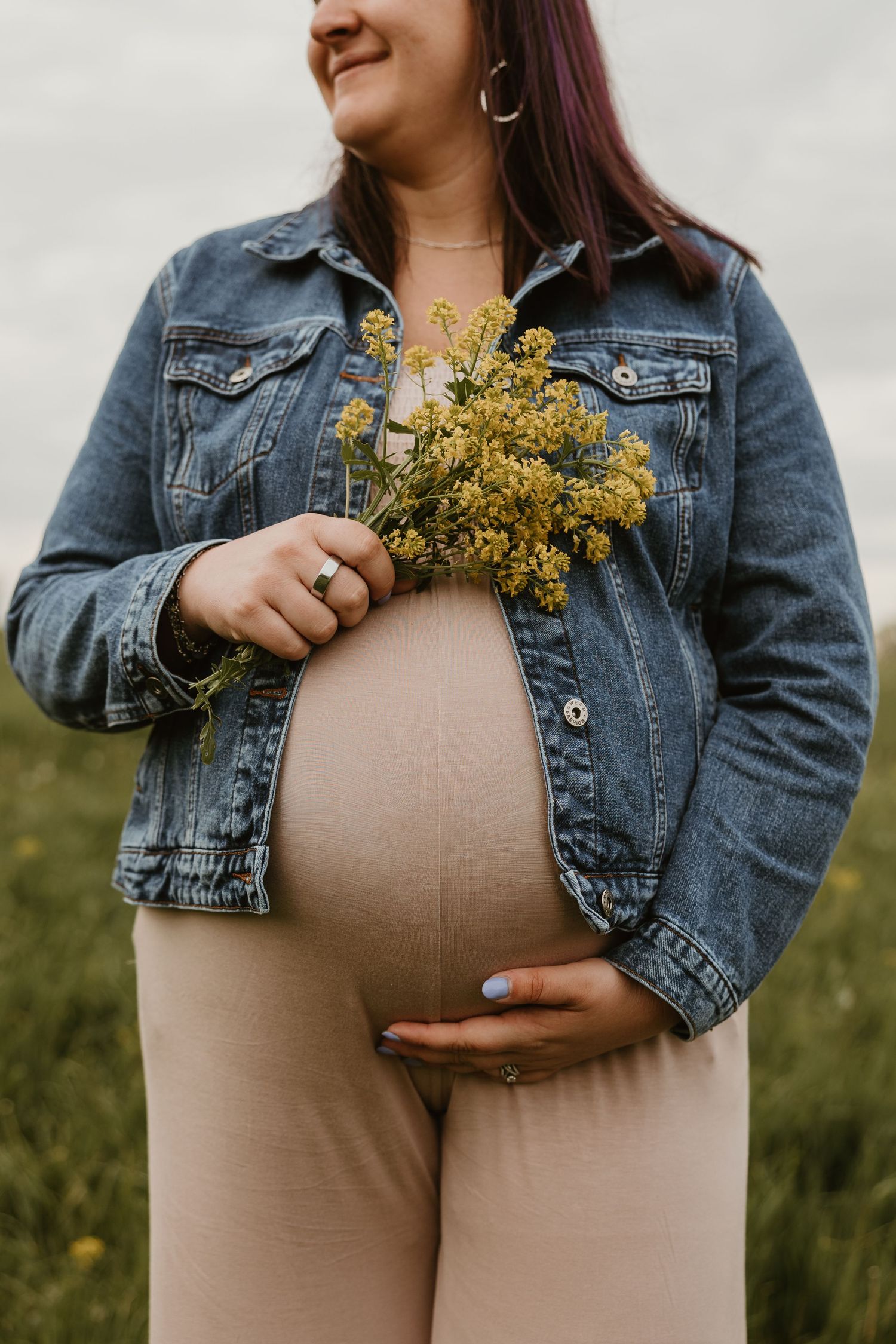 Pregnant woman in denim jacket holding wildflowers, cradling baby bump outdoors in a field.