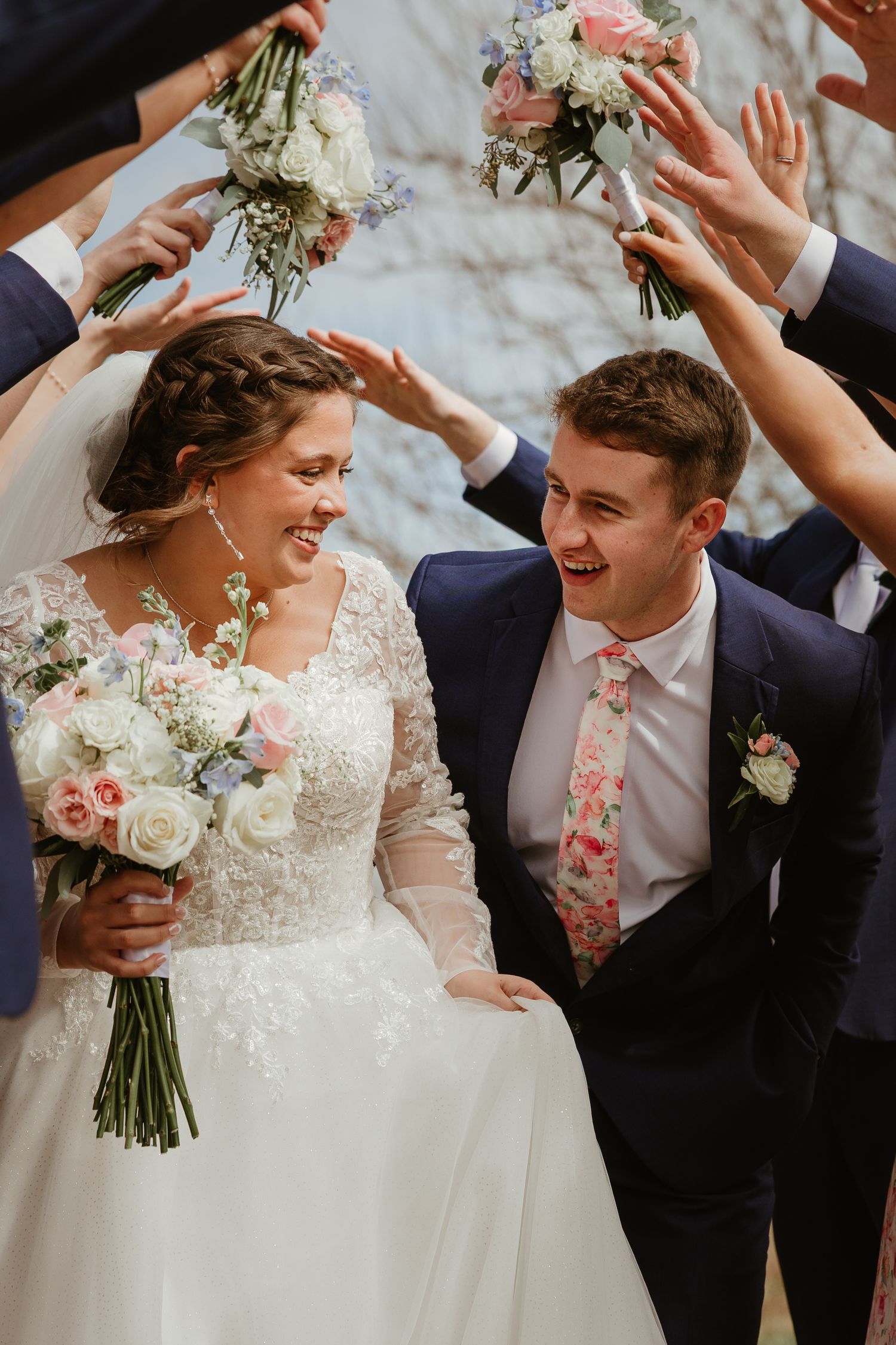 Joyful bride and groom laughing as wedding party raises bouquets overhead in a celebratory arch.