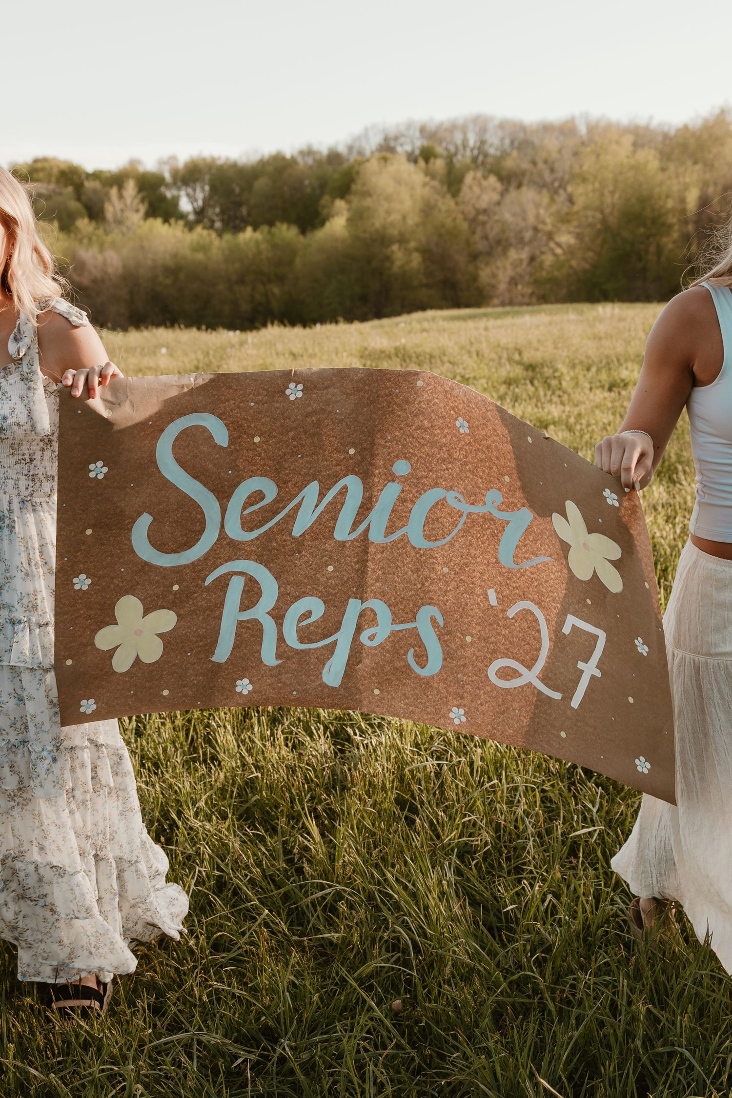 Two girls hold a decorated cardboard sign reading Senior Reps 27 in a green field.