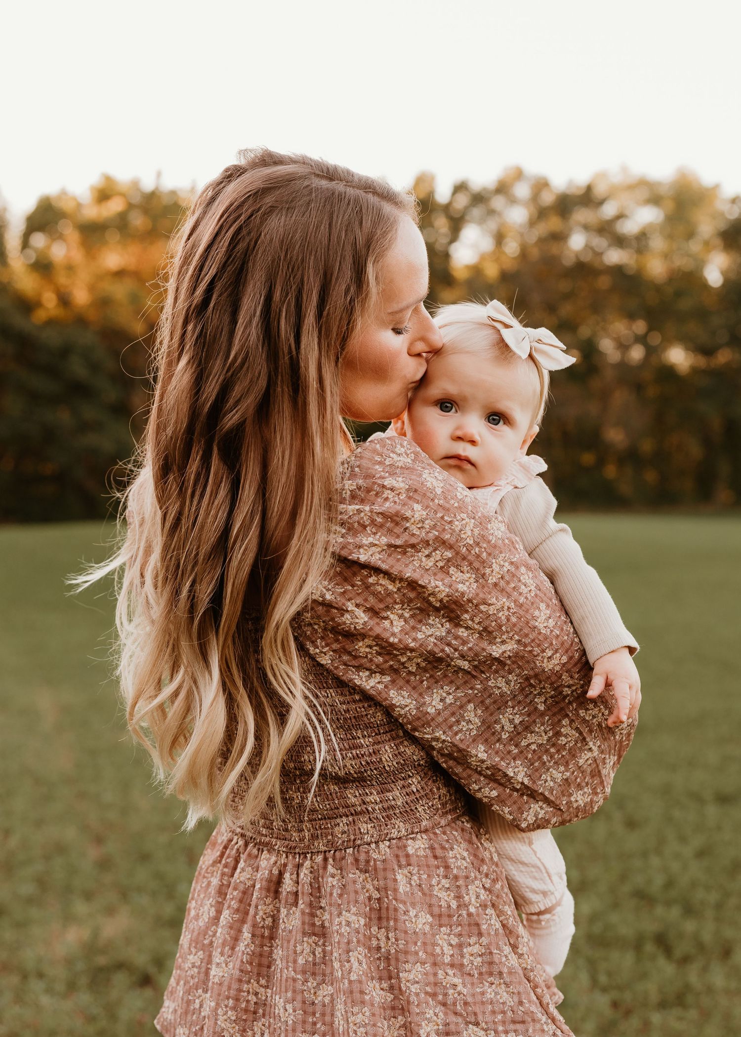A mother tenderly kisses her baby girl outdoors in a sunlit field, both wearing soft floral outfits.