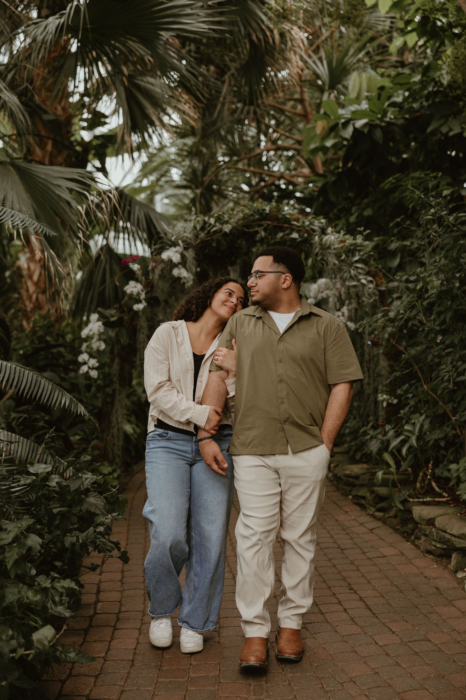 Couple walking hand in hand through a lush tropical greenhouse pathway, smiling at each other.