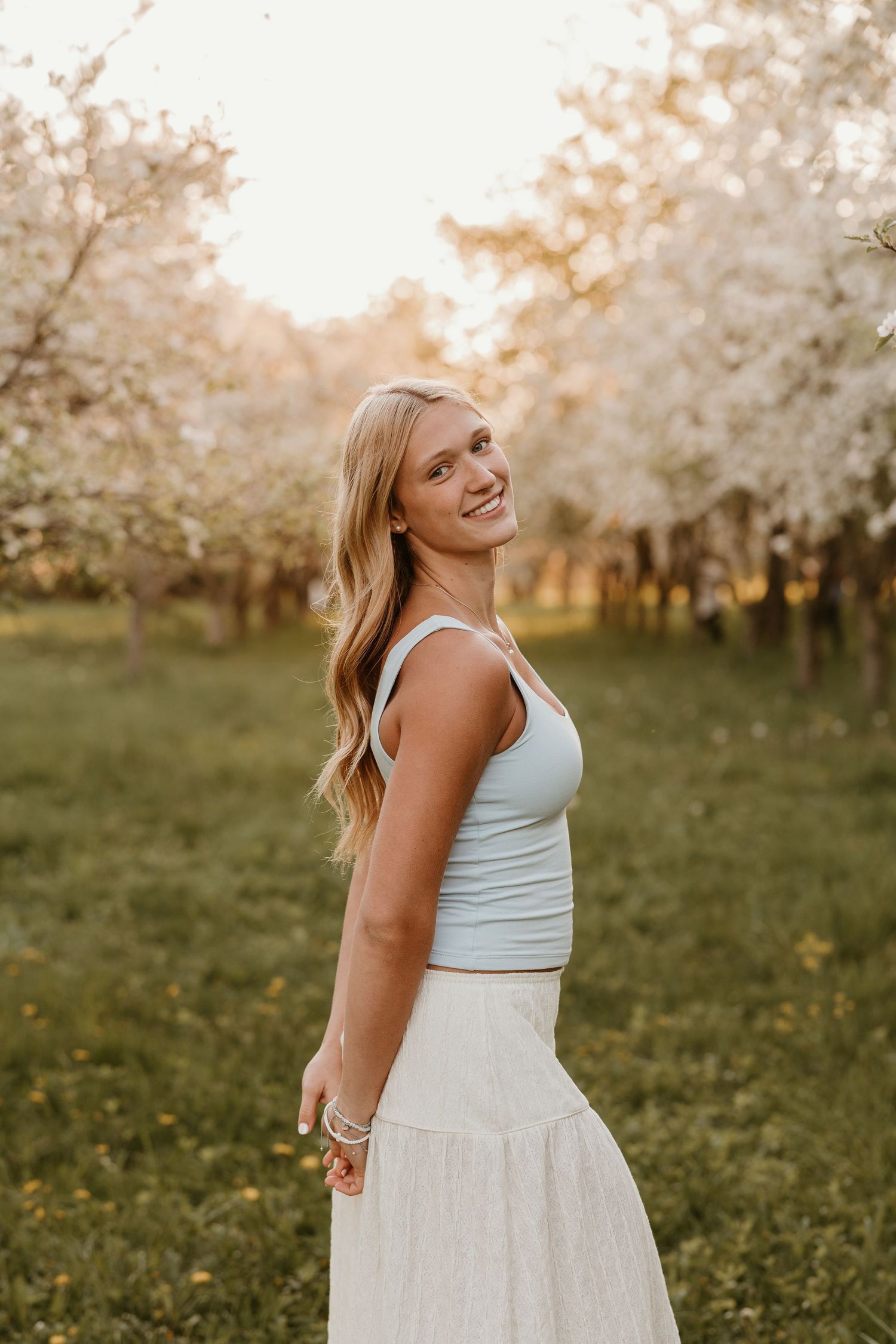 Young blonde woman smiling in white skirt and light blue top among blooming spring trees at golden hour.