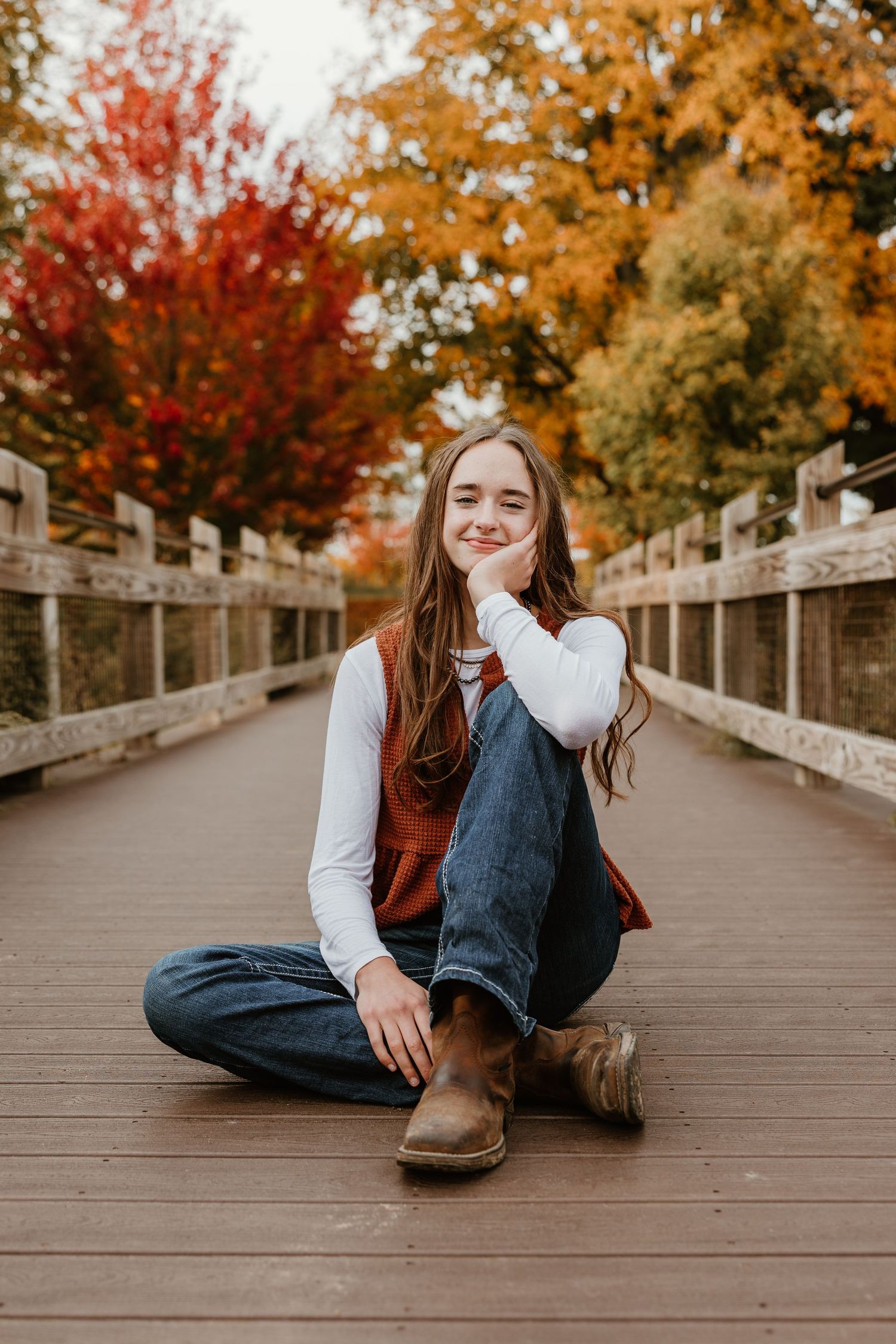 Young woman sitting cross-legged on wooden bridge surrounded by vibrant autumn foliage in red, orange, and gold.