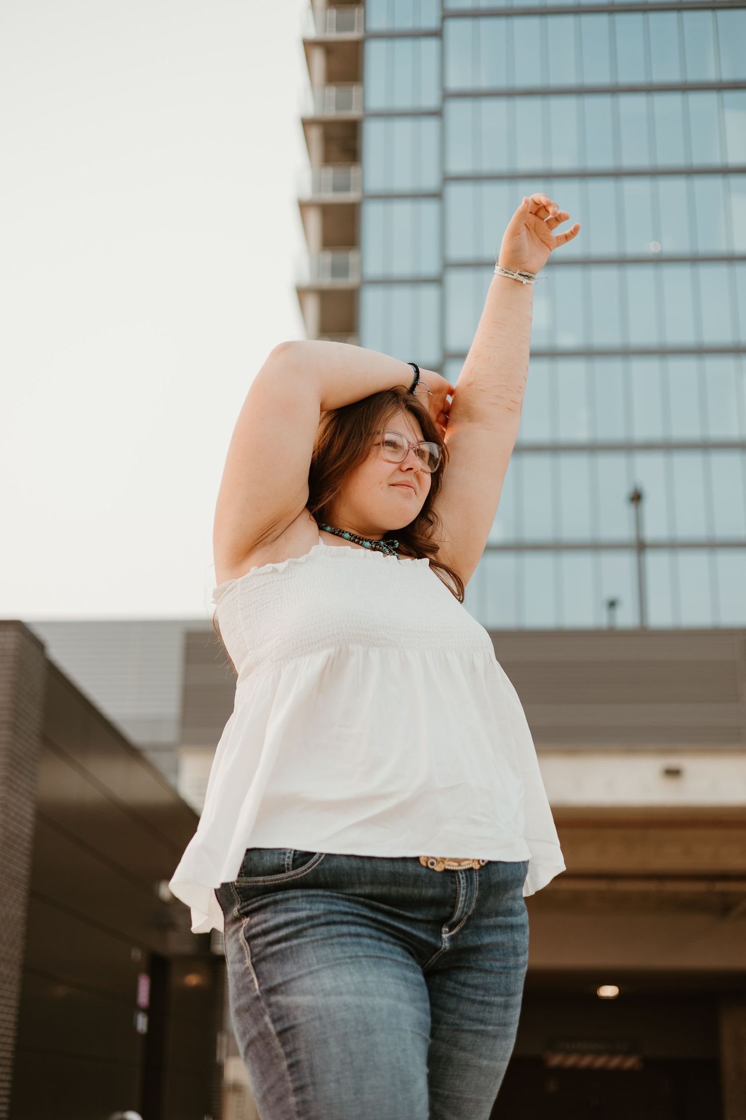Plus-size woman in white top and jeans posing outdoors with arms raised near modern building.