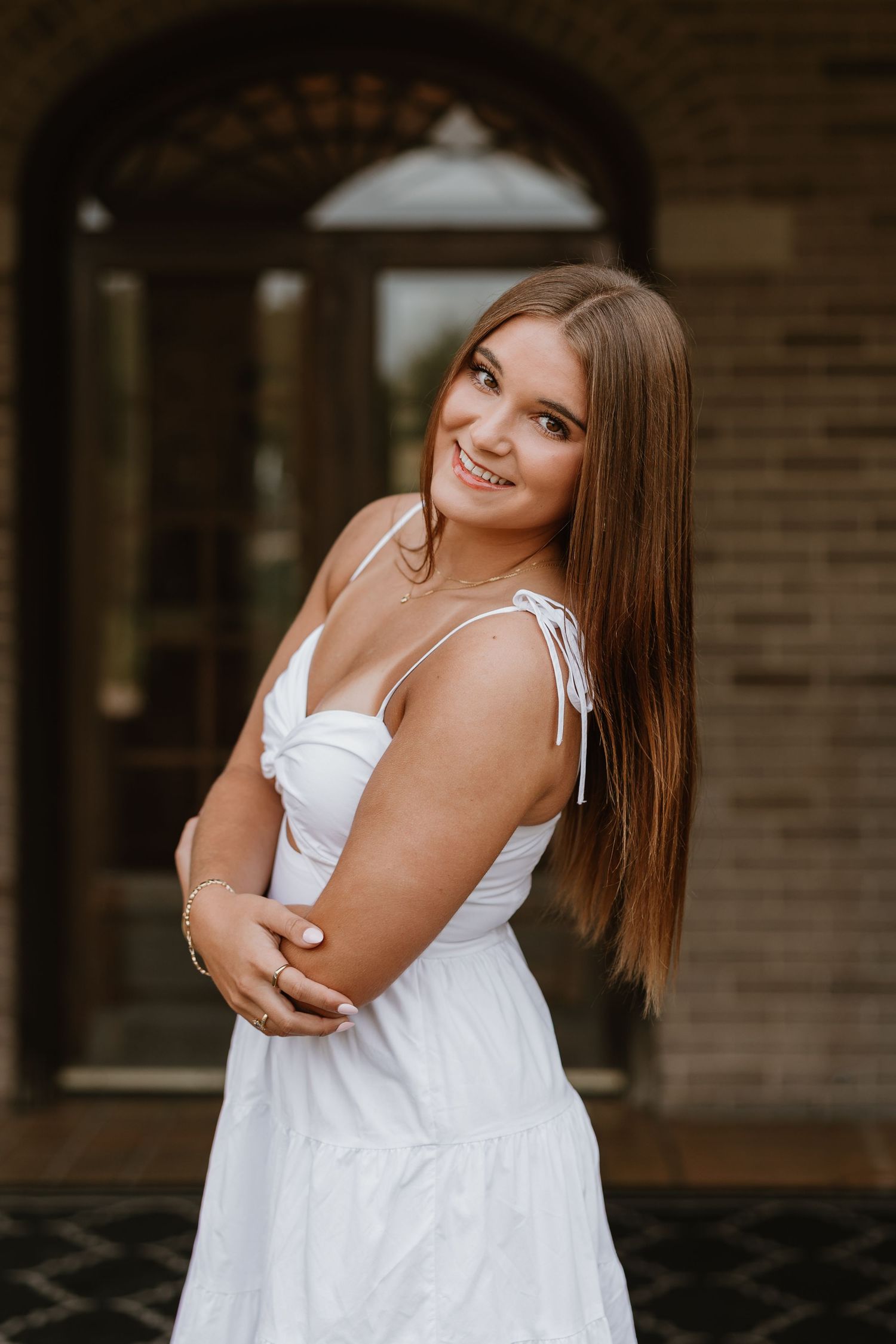Young woman in white sundress smiling over shoulder, posing outdoors near brick building with arched doorway.