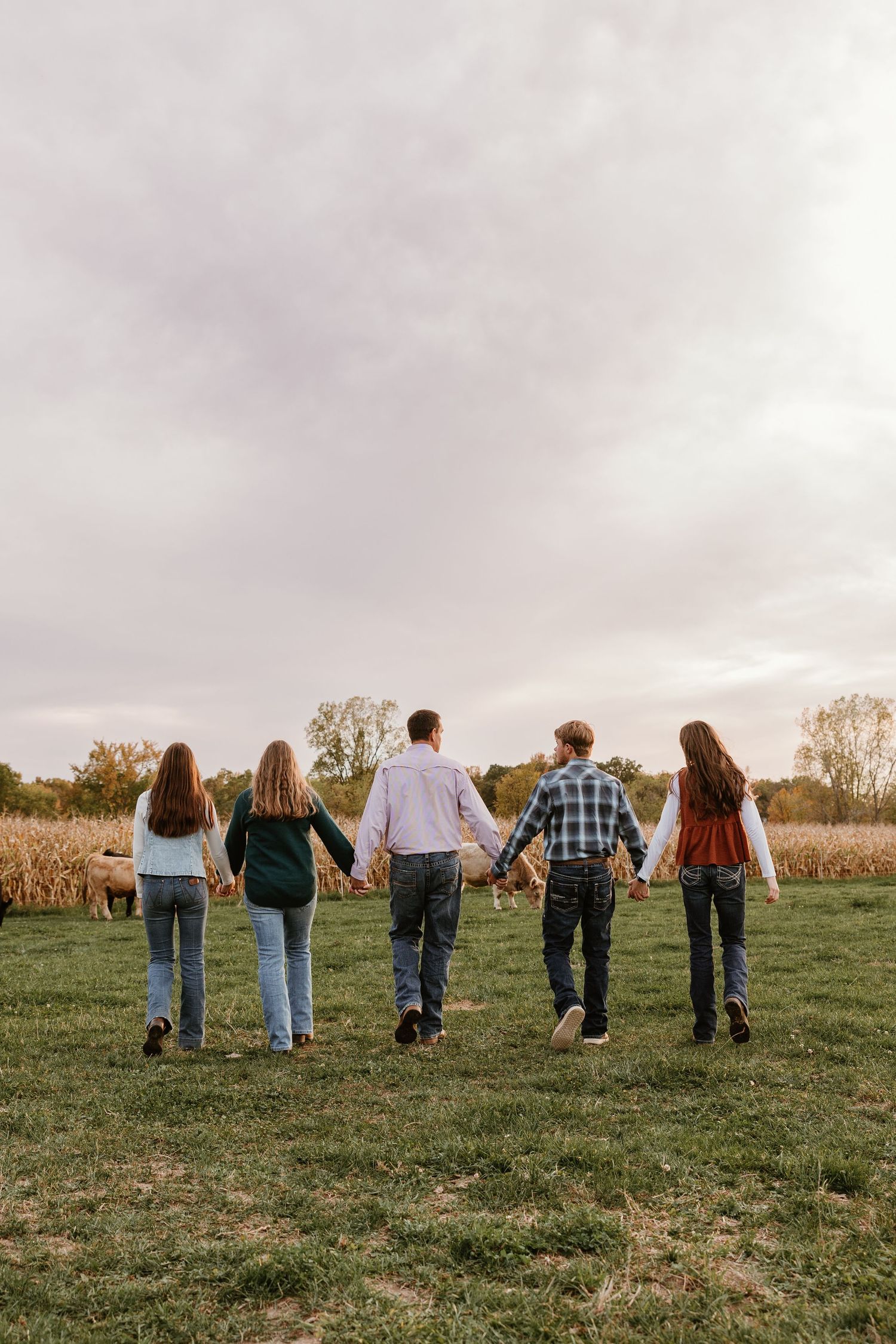 Five people holding hands walk through a green field near a cornfield on a cloudy autumn day.
