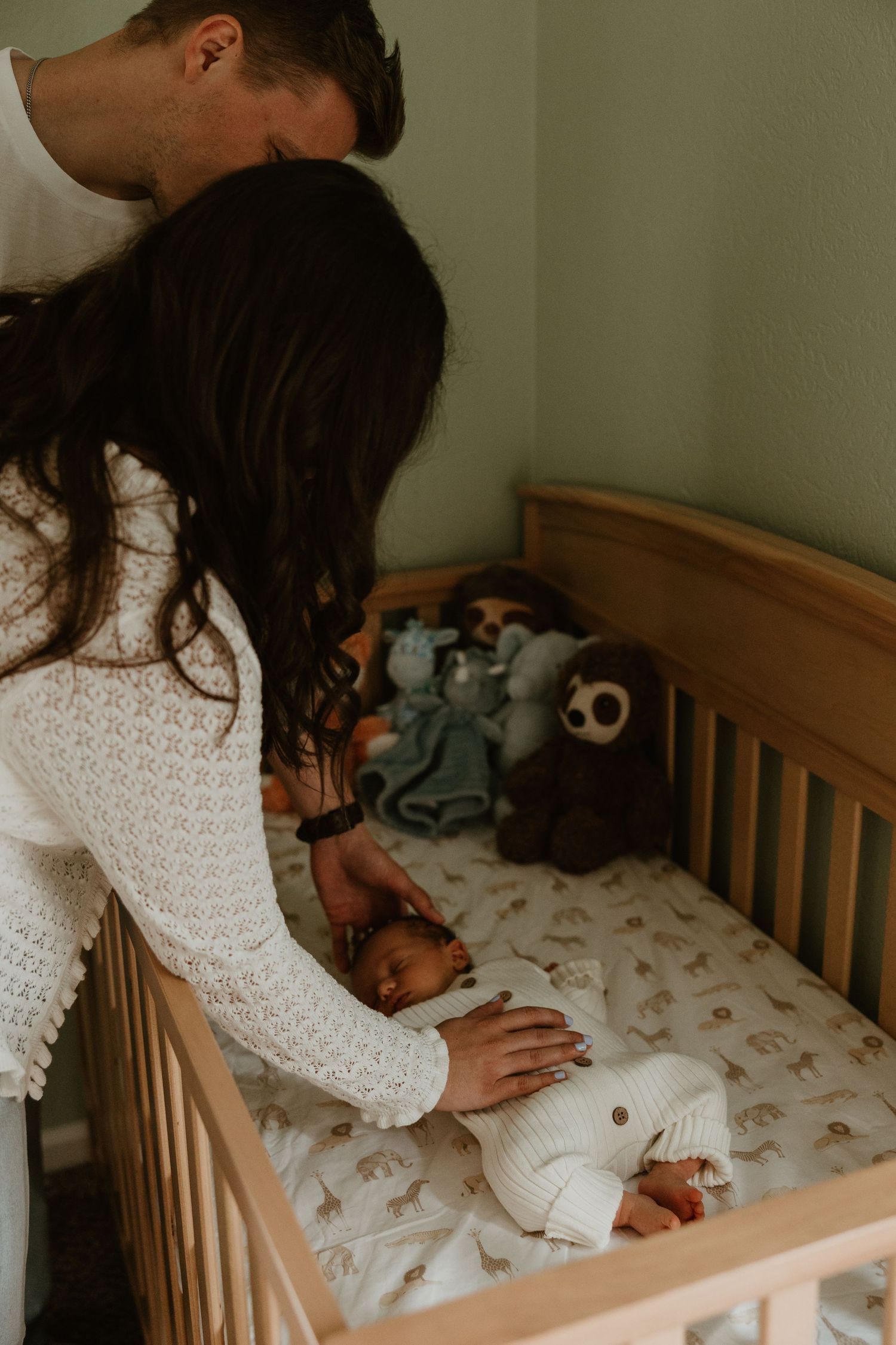 Parents lovingly place their newborn baby in a wooden crib with stuffed animals in a sage green nursery.