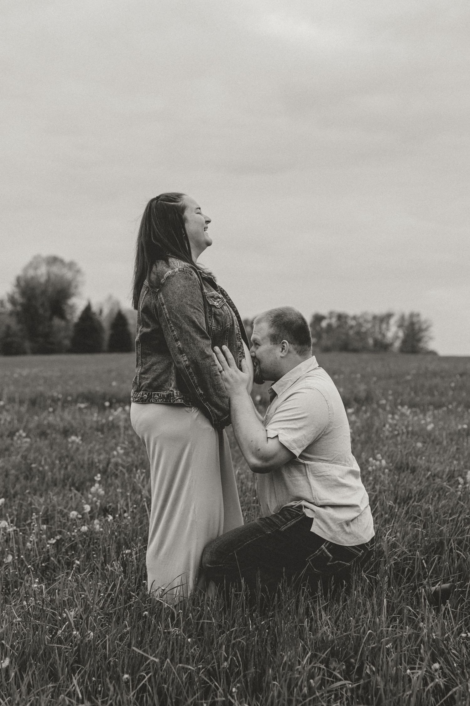 Black and white maternity photo of man kneeling in field, kissing pregnant woman's belly outdoors.