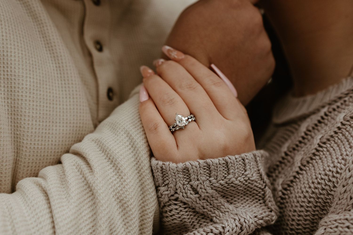 Close-up of a woman's hand wearing a diamond engagement ring, resting on a man's arm in cozy sweaters.