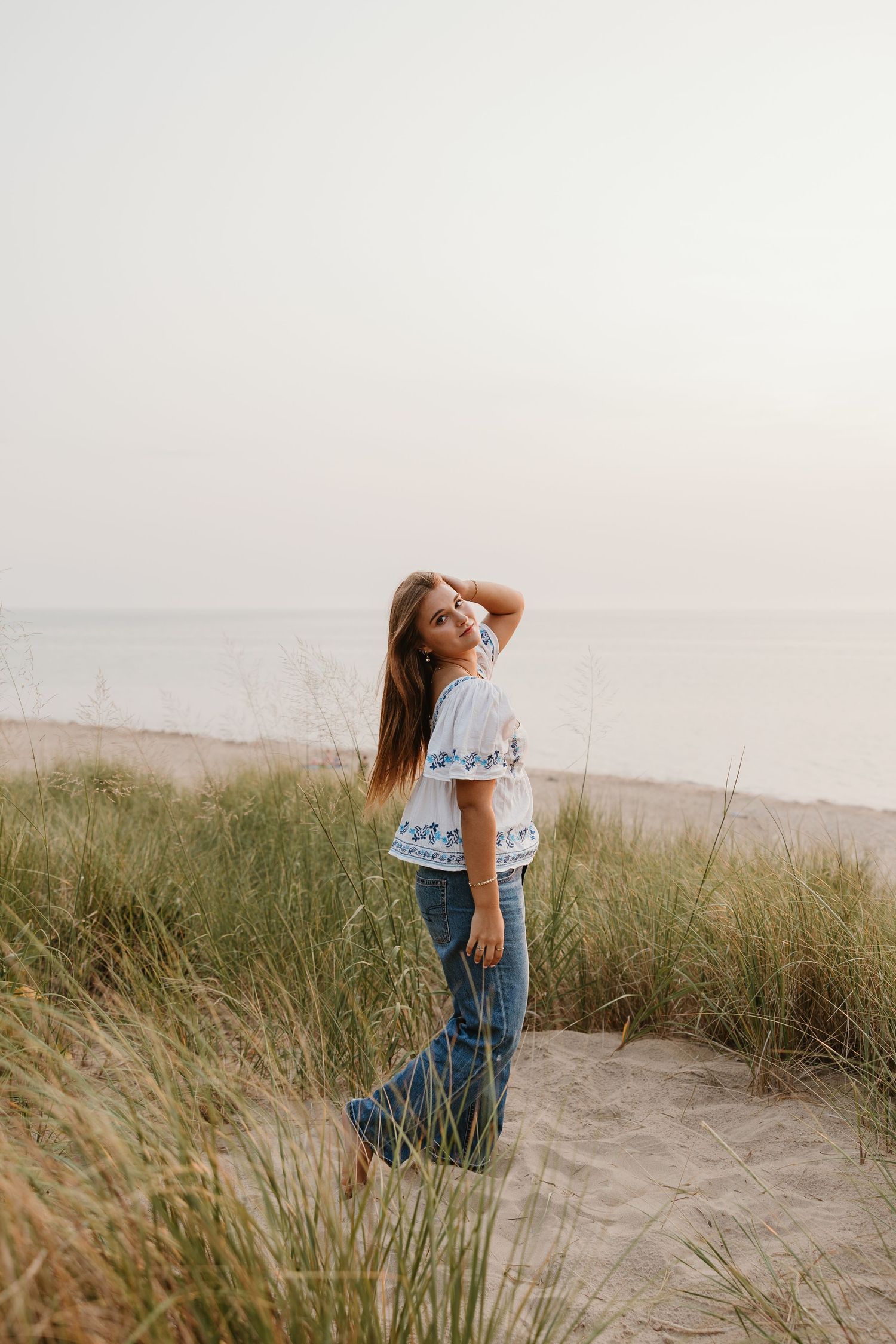 Young woman in white embroidered top and jeans poses on sandy beach dunes with tall grass and ocean backdrop.